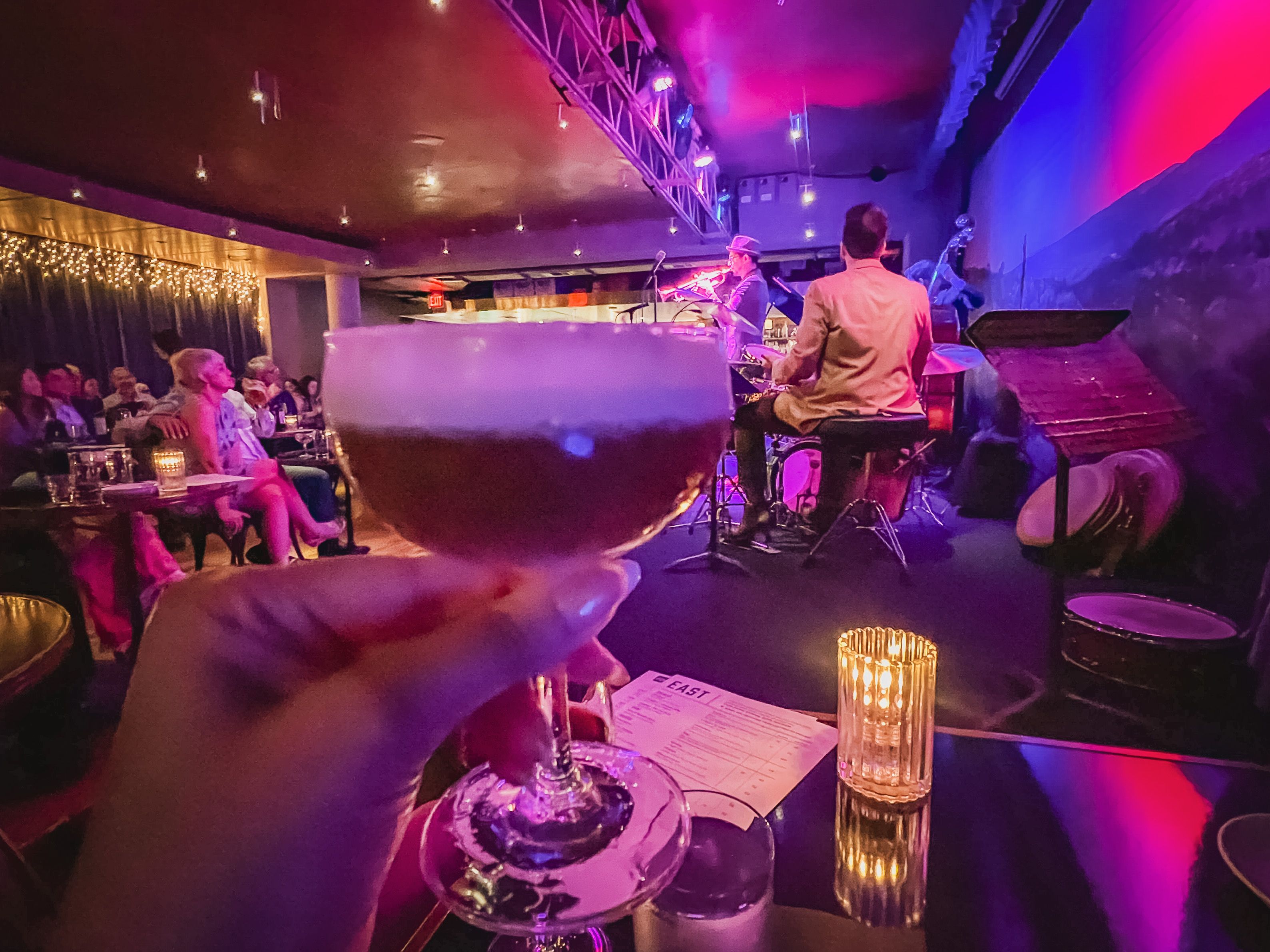 A dark-colored cocktail with a foam layer with a jazz band in the background. 