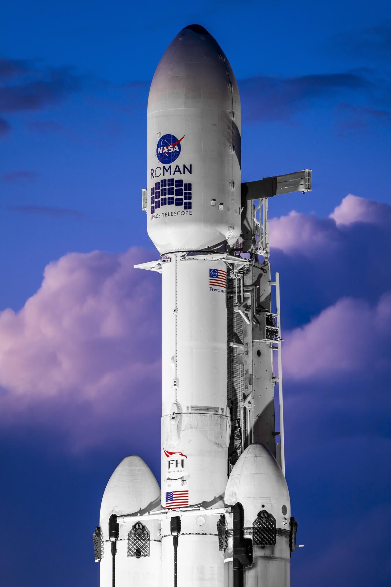 Tall white rocket on a launch pad against a deep blue evening sky with purple clouds. NASA logo and ROMAN branding along with Space Telescope text visible, plus a support tower and American flag.