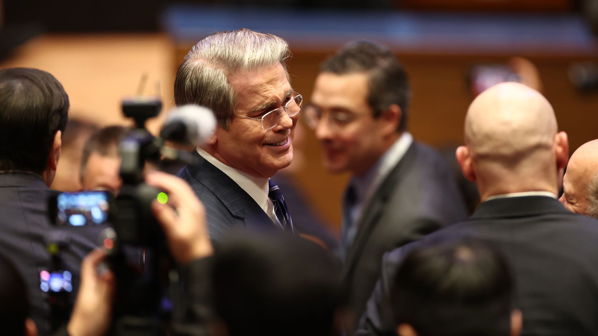 Treasury Secretary Scott Bessent, in a suit and glasses, surrounded by people and cameras at an event. 