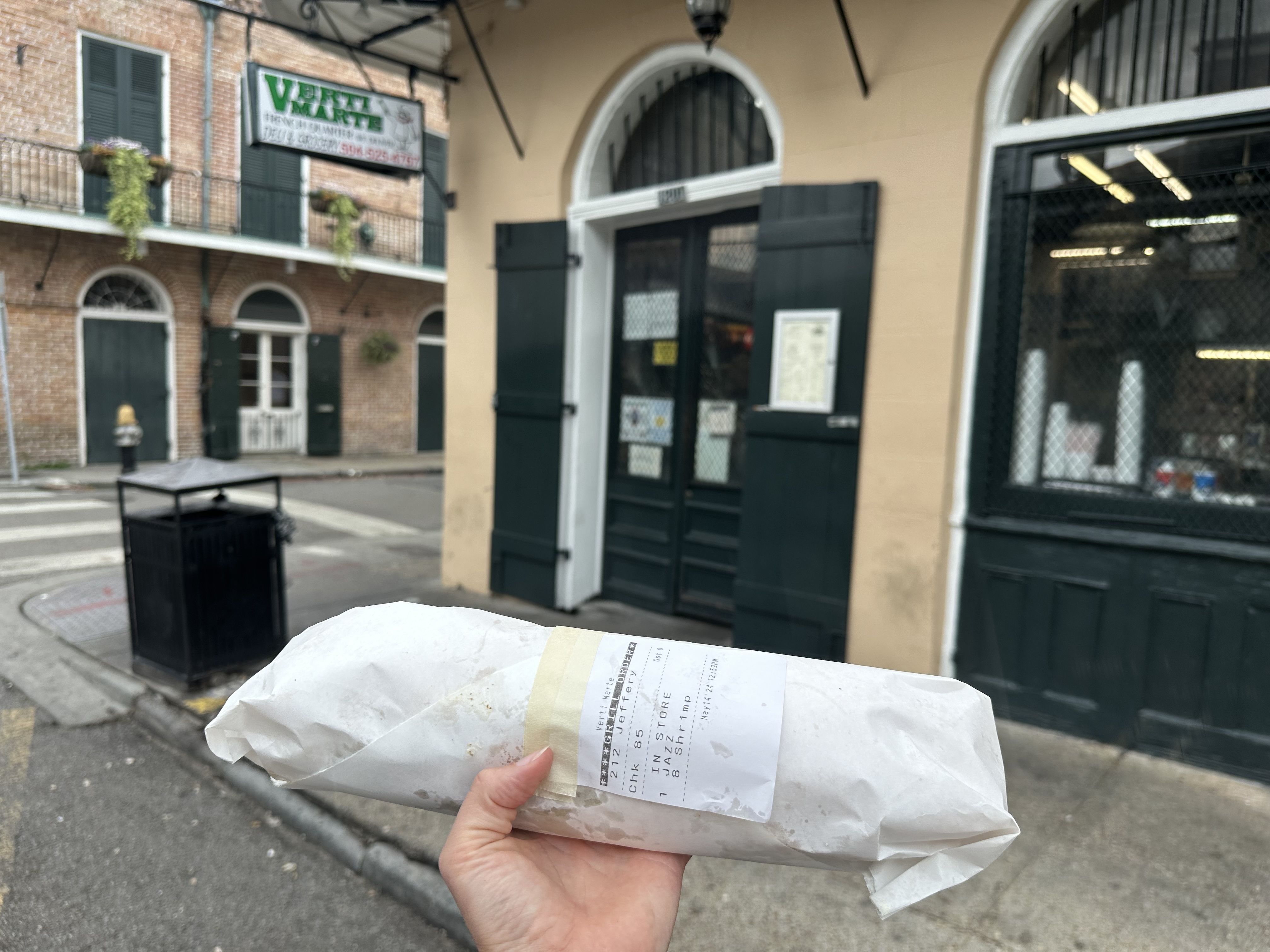 A photo of a hand holding up a butcher paper-wrapped poboy in front of a corner store. The store sign says Verti Marte.