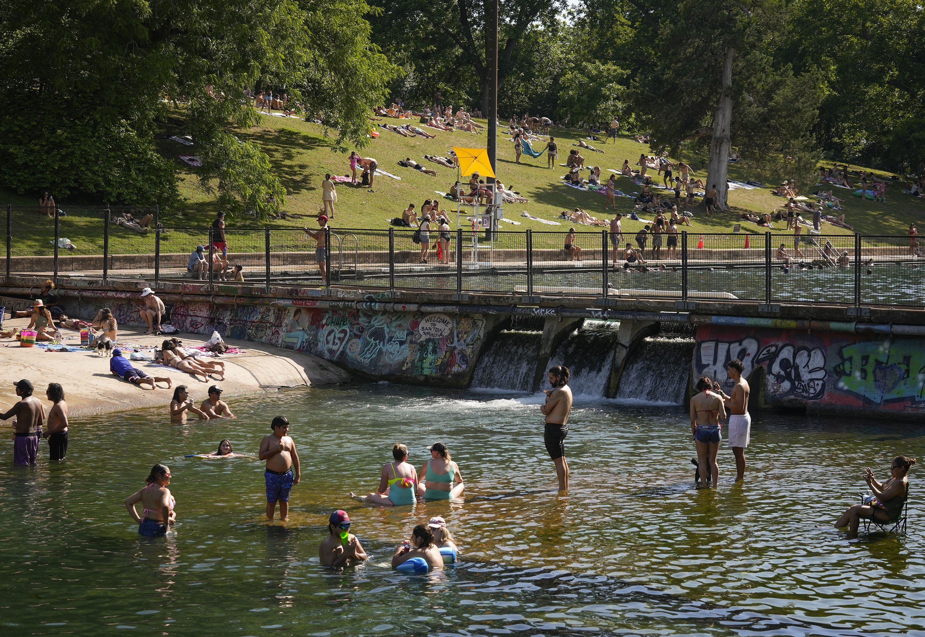 People soak up sun on a grassy riverbank under leafy trees; a yellow canopy shades a lifeguard chair. Many swimmers in the water; graffiti-covered canal walls line the foreground behind a fence.
