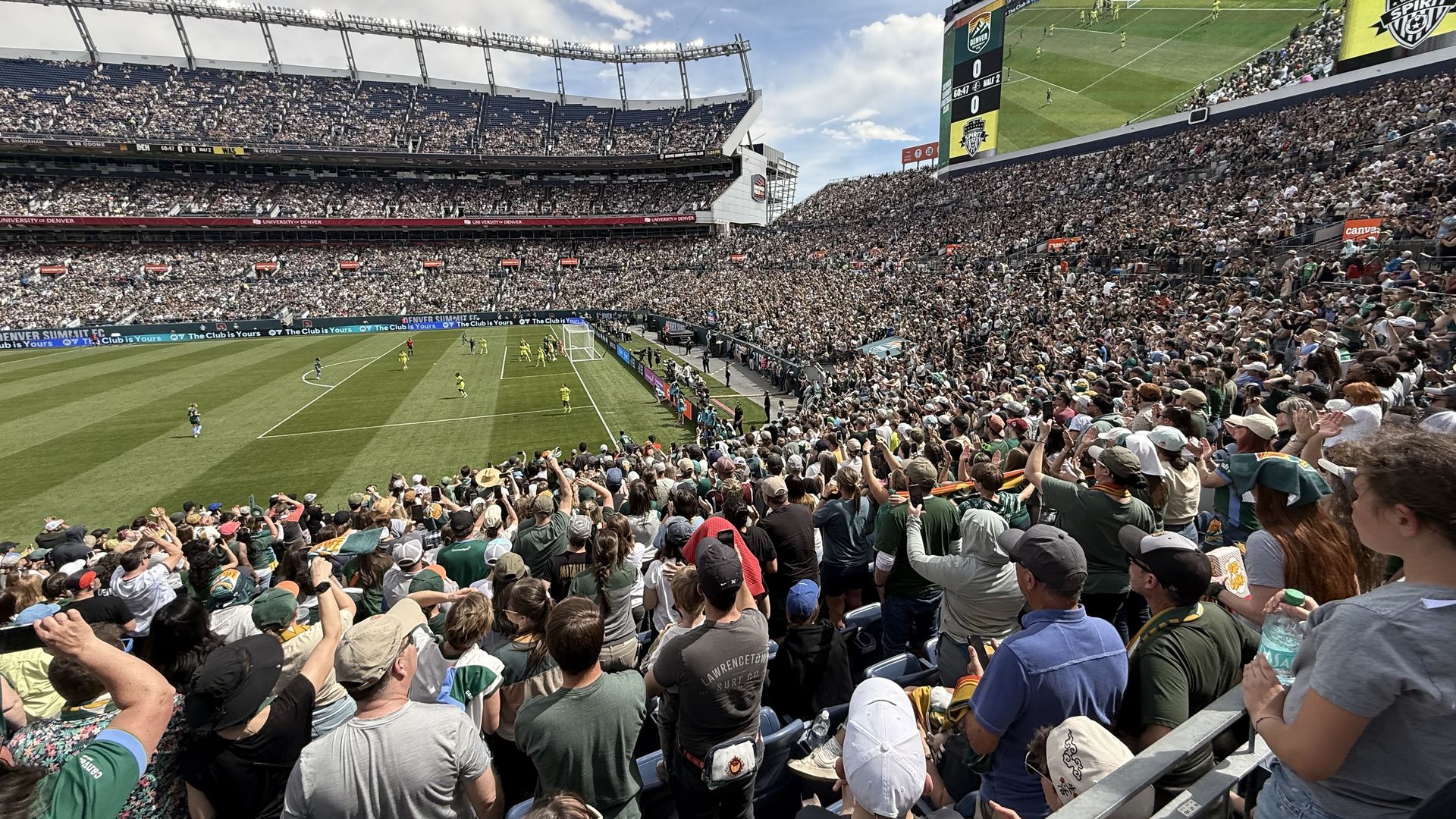 Panoramic view of a packed outdoor soccer stadium during a match; bright green field, players near the goal, and a sea of fans in the stands beneath a large video scoreboard.