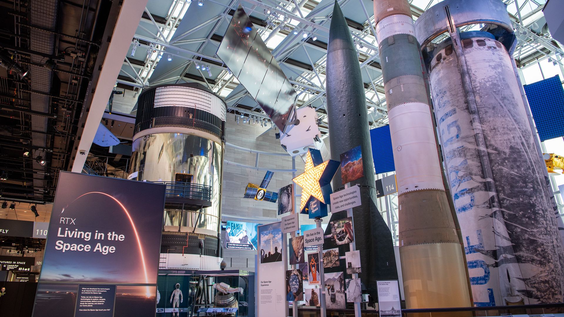 Indoor space museum exhibit featuring tall silver rocket models, cylindrical boosters, and display panels under a glass and steel roof.