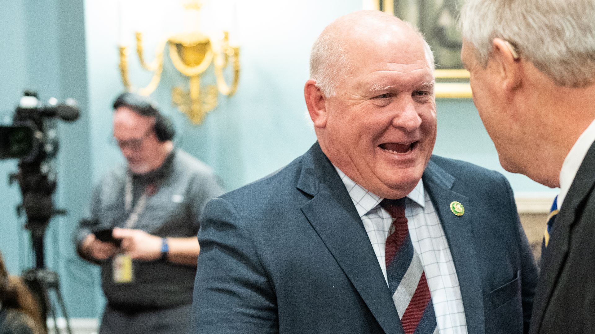 Two older men shaking hands in a government space. Both in suits. The face of only one can be seen.