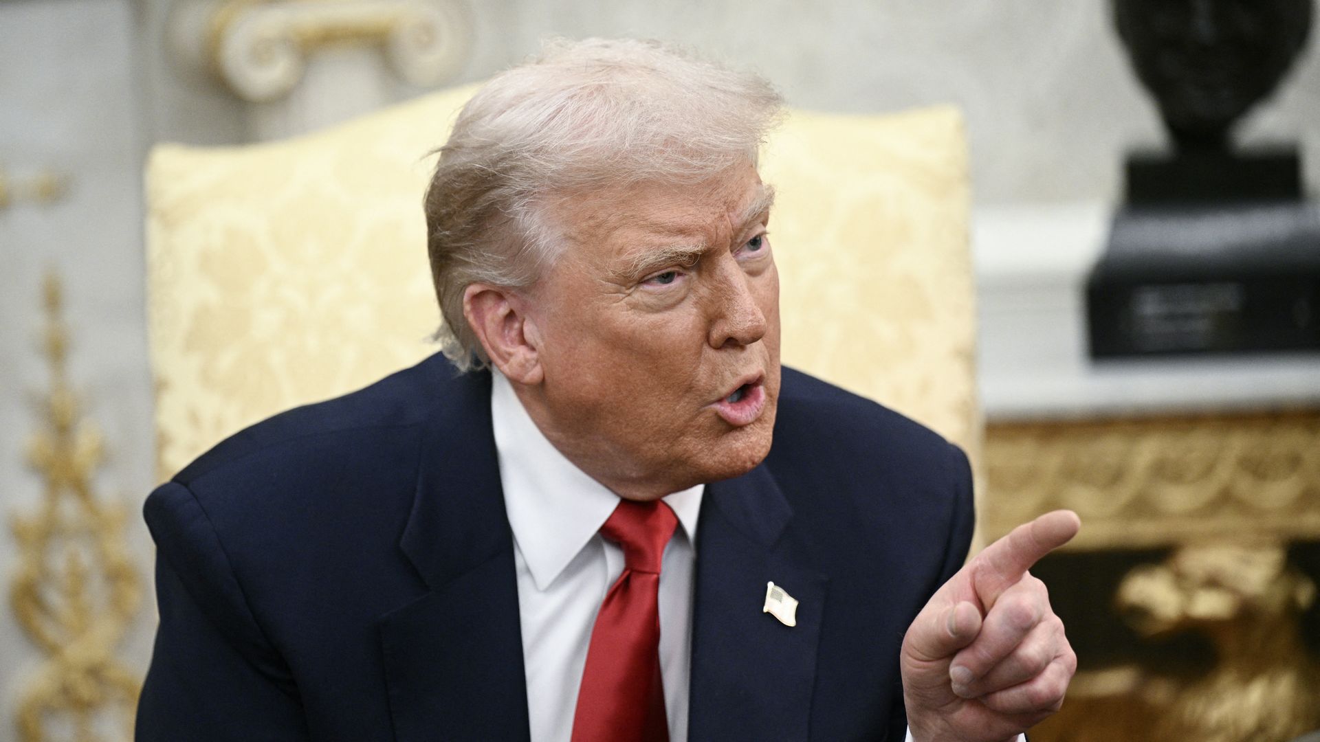 President Trump wearing a blue suit and pointing out of frame while sitting on a gold chair in a white room.