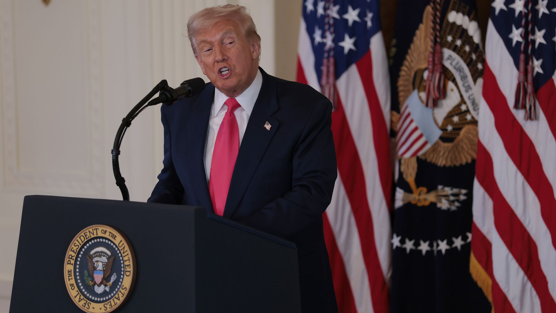 President Trump, wearing a navy jacket with a US flag pin at the top of his left lapel, white shirt and red tie, looks to his left as he speaks at a podium.