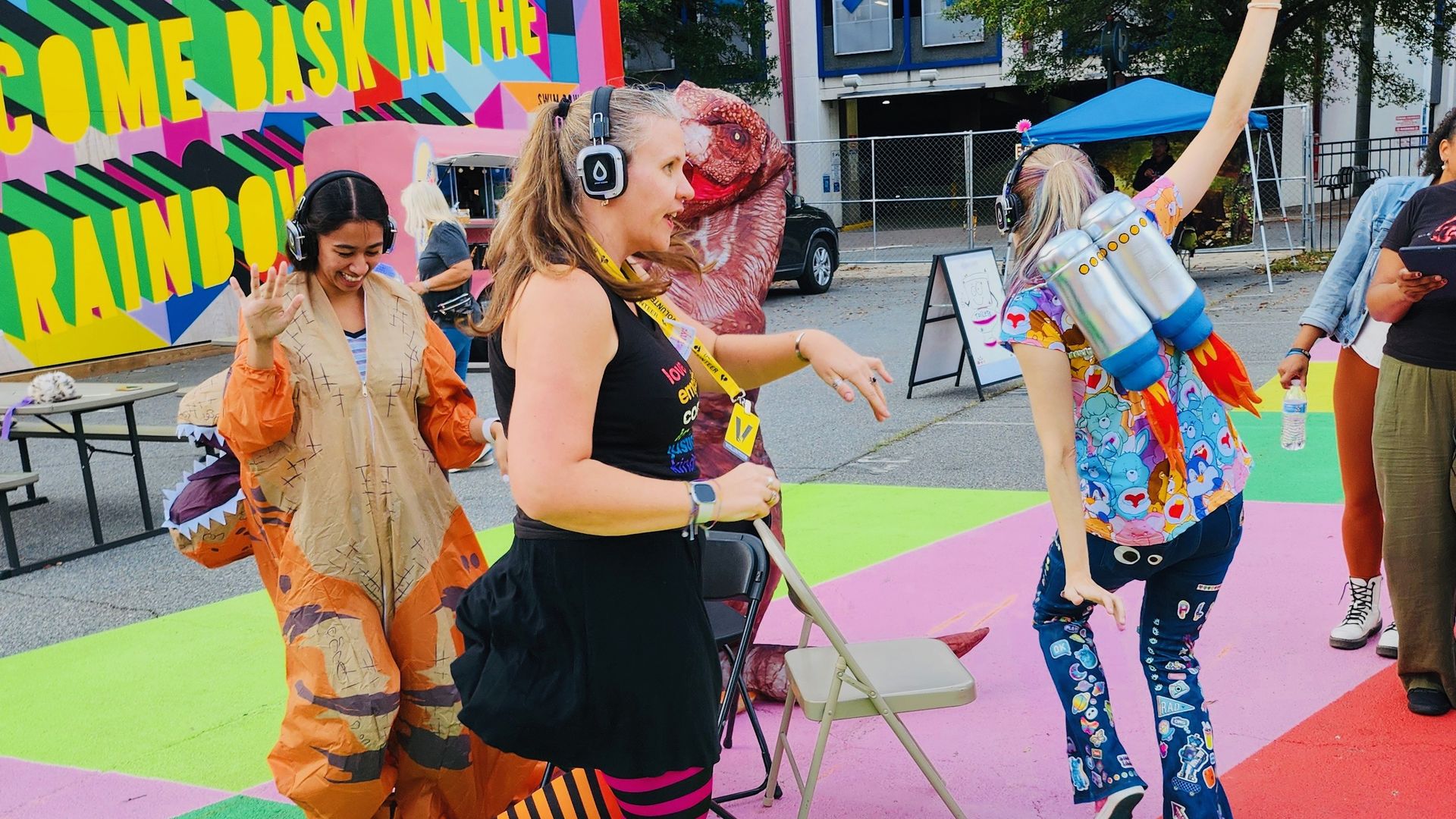 People dancing outdoors on colorful geometric pavement, wearing headphones, one in a dinosaur costume, another with a jetpack prop, in front of a bright mural reading "COME BASK IN THE RAINBOW".