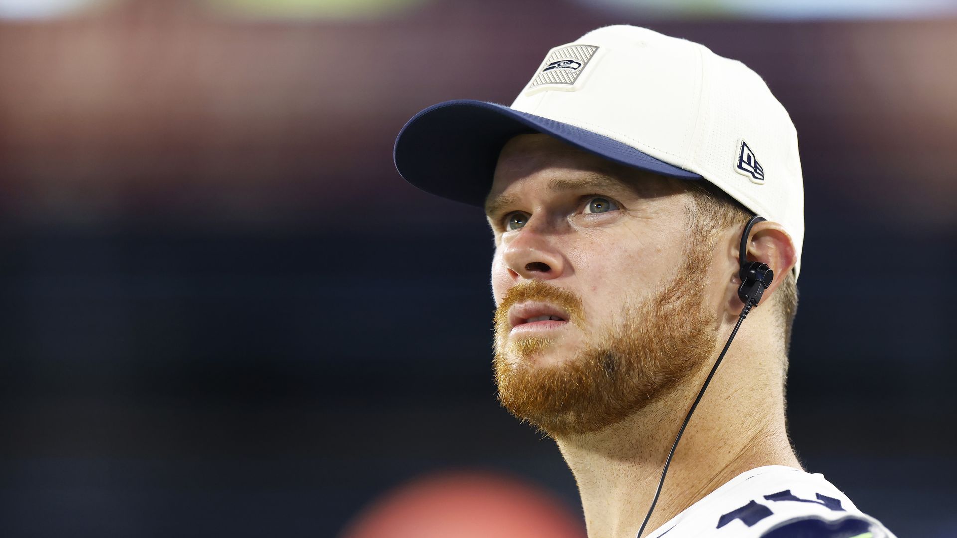 Seahawks quarterback Sam Darnold looks upward while wearing a headset and Seahawks cap.