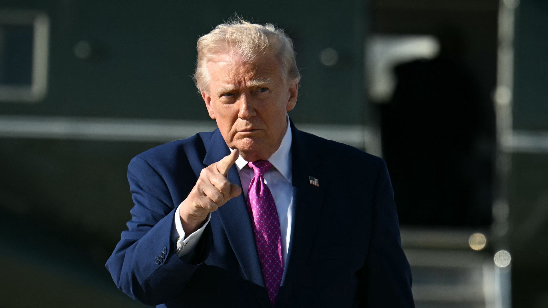 A man in a navy suit and bright pink tie points a finger at the camera outdoors, with a small American flag pin on his lapel and a serious, focused expression.