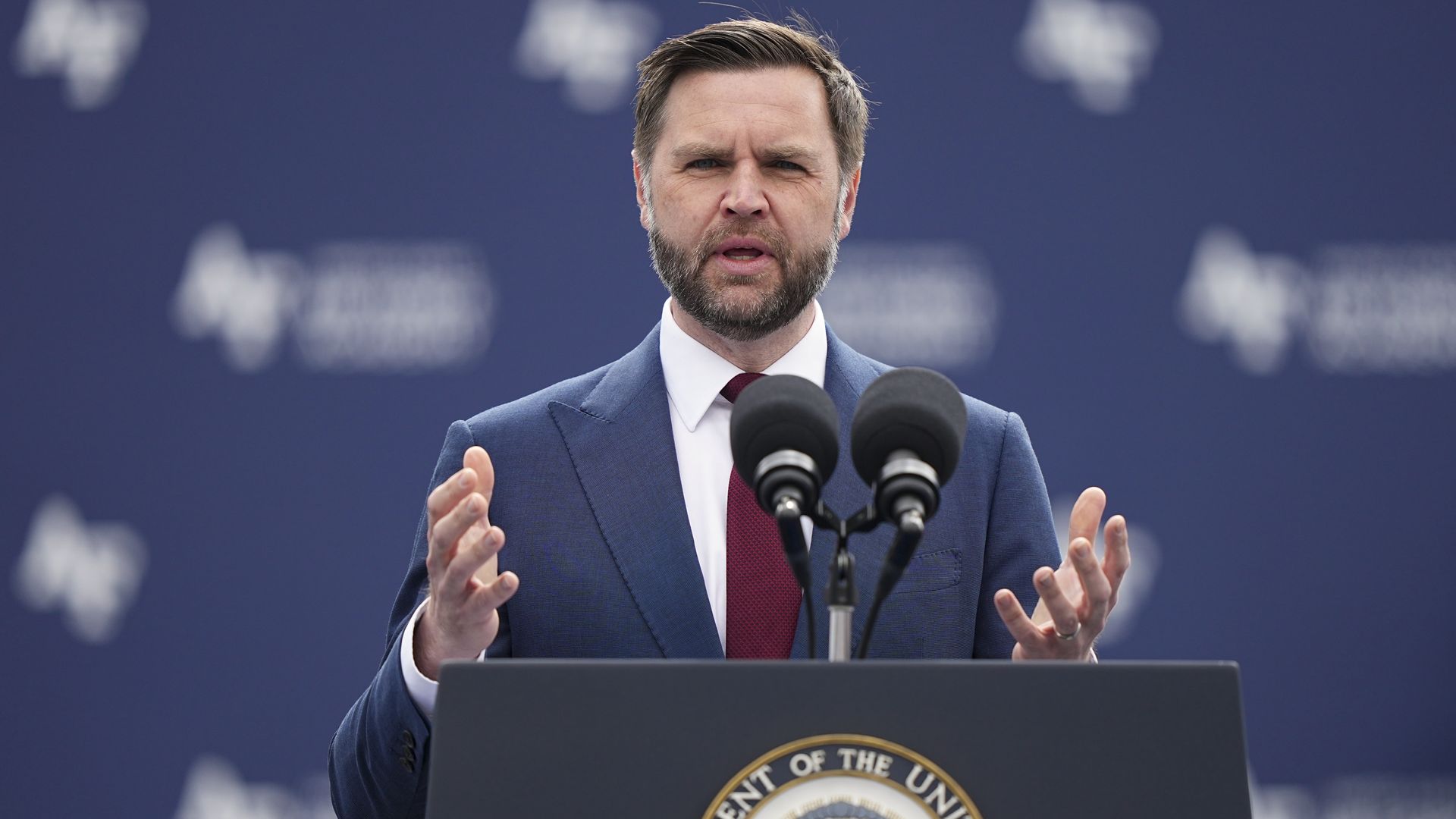 Vice President JD Vance, a bearded man in a blue suit and red tie speaks at a podium with the presidential seal, flanked by microphones, against a dark blue backdrop.