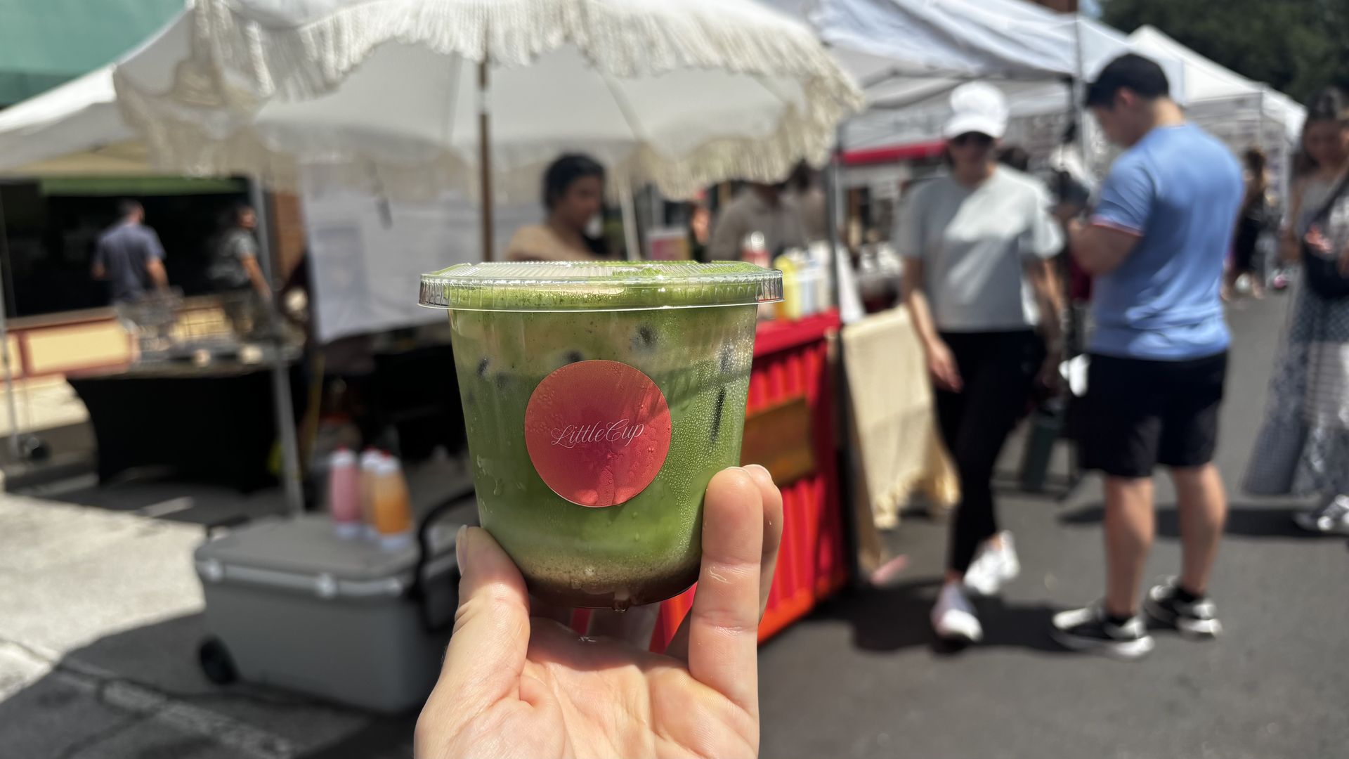 Close-up of a green smoothie in a clear cup with a pink label "LittleCup" on it, held by a person. Outdoor market stalls and people blur in the background.