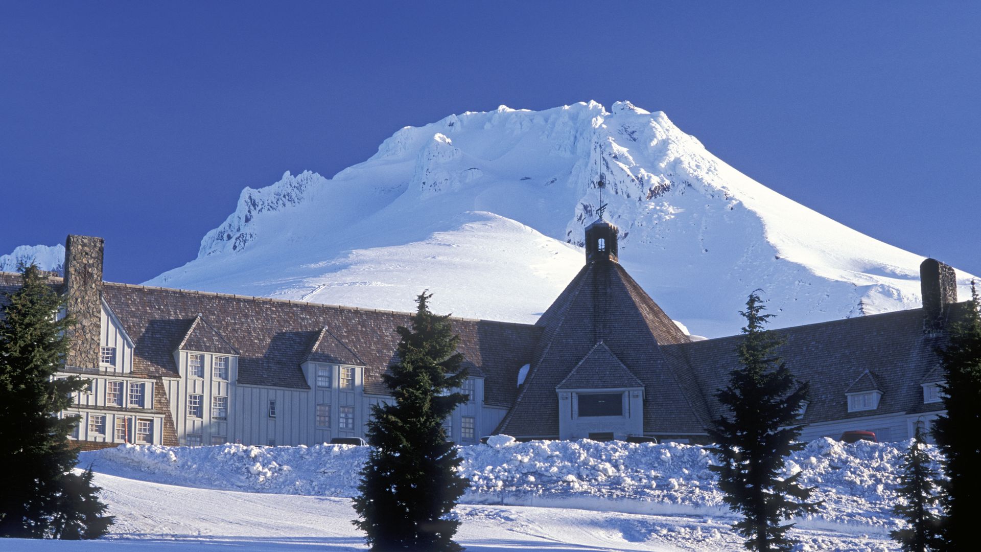 A photo of Mount Hood in winter shows the Timberline lodge in the foreground with the summit sits covered in snow above the building.