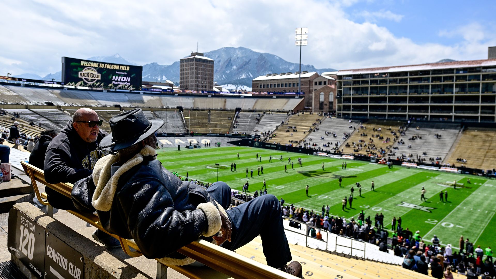 Two fans sit in an empty seating section under the sun at Folsom Field as players warm up on the field.