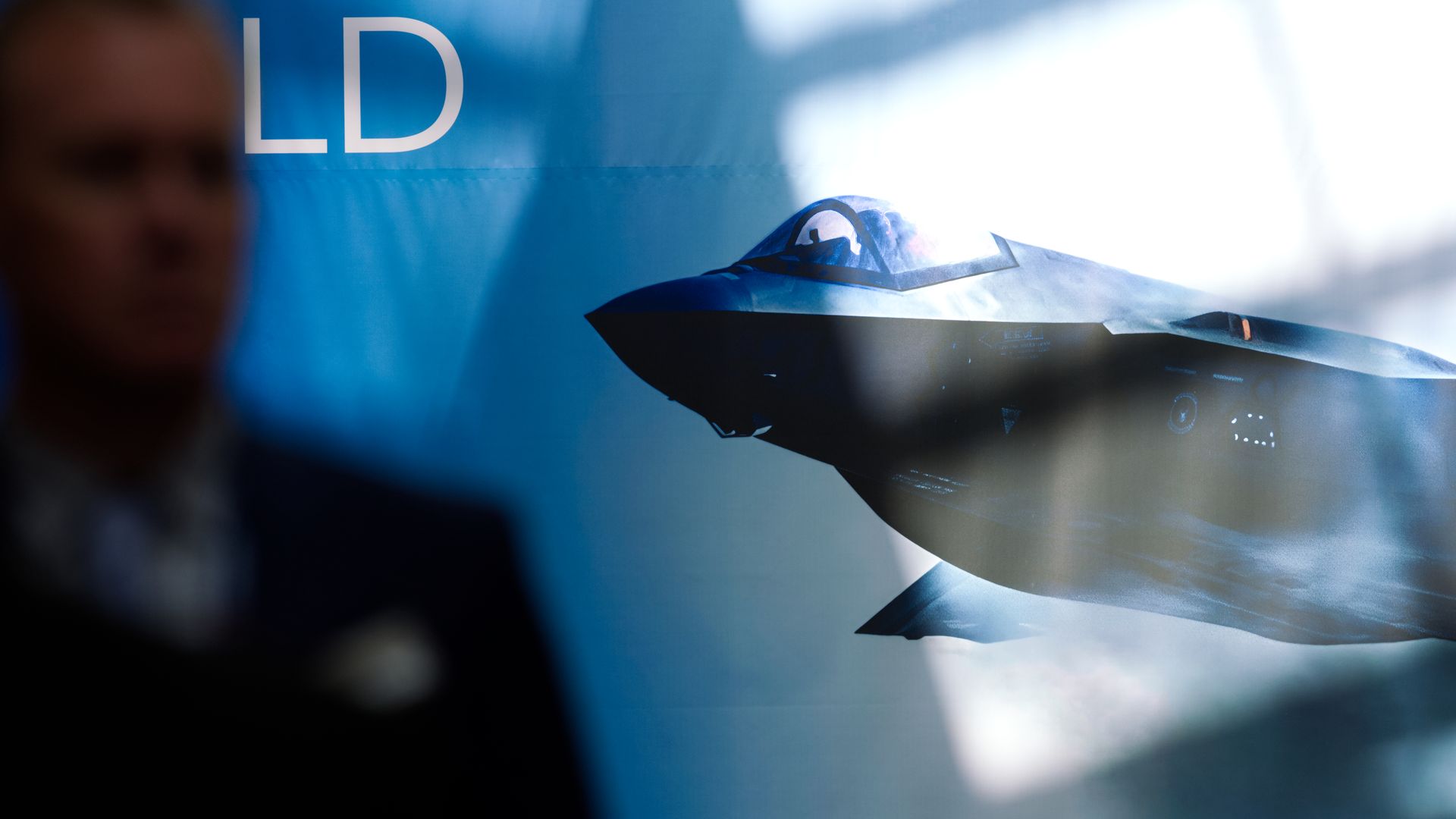 Blurred silhouette of a man in a suit on the left with a detailed image of a stealth fighter jet on a blue background to the right.