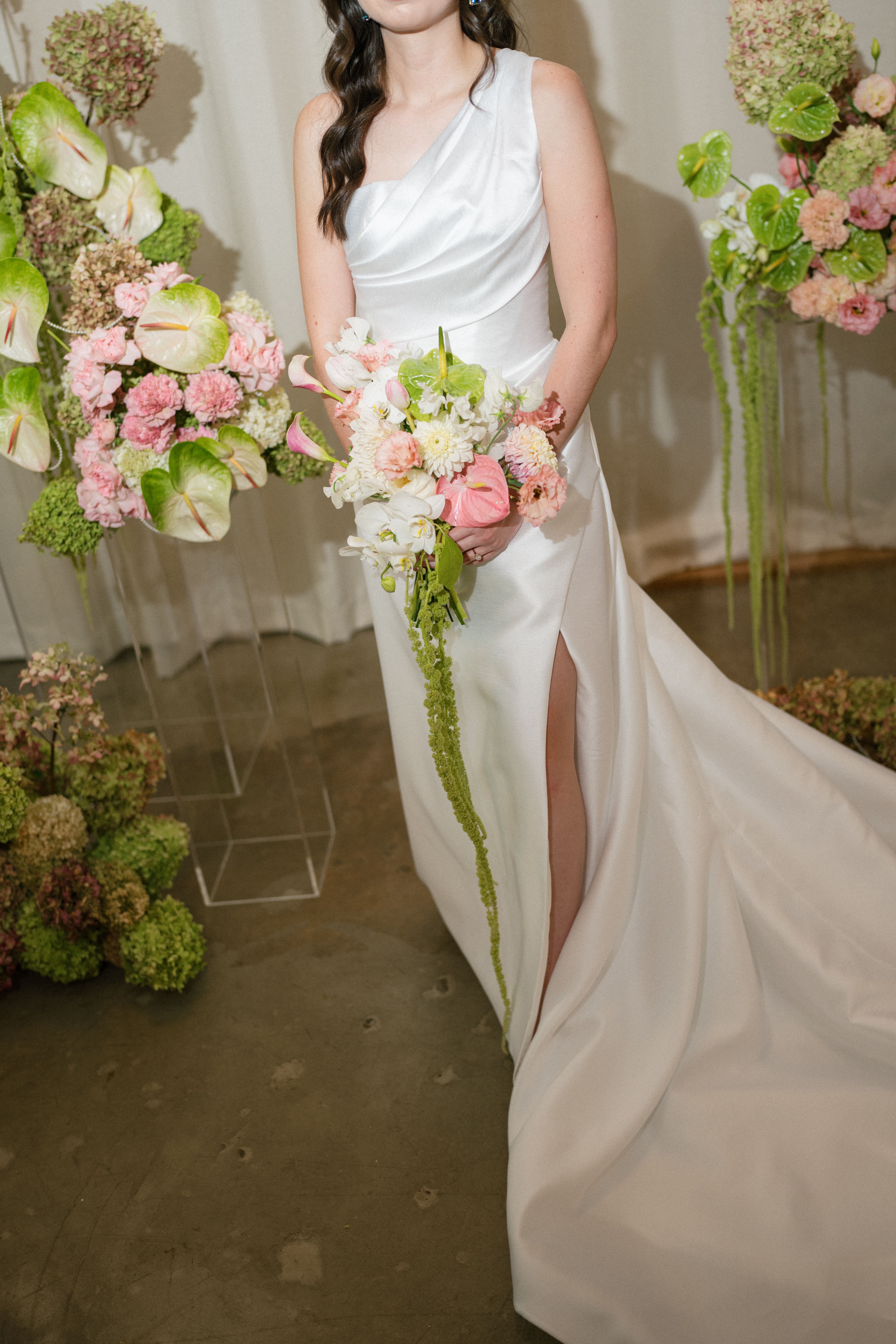 Woman in a white one-shoulder wedding gown with a thigh slit holding a bouquet of pink, white, and green flowers, standing among floral arrangements with similar colors.