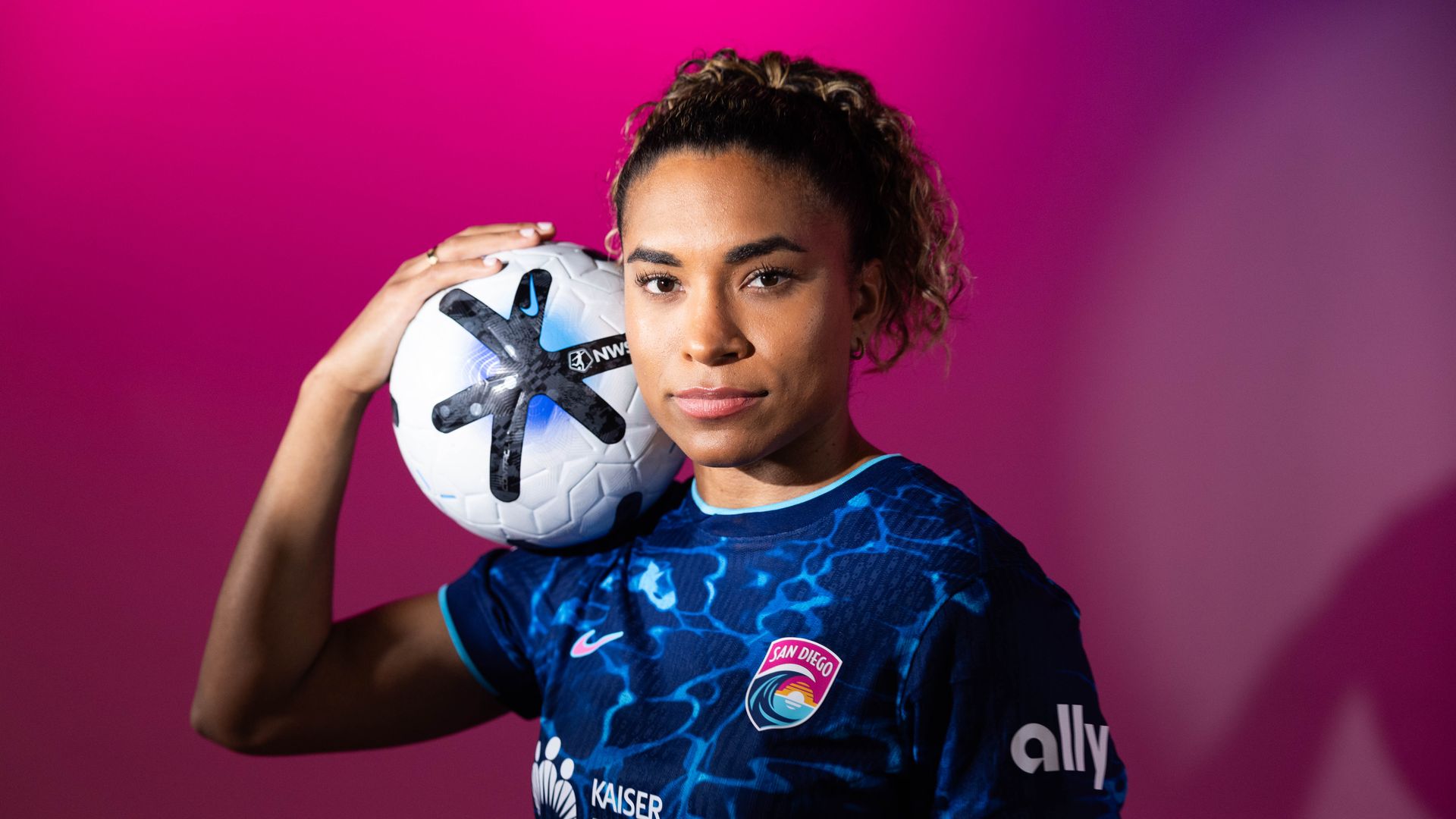 Portrait of a female soccer player in a blue San Diego crest jersey, holding a white soccer ball against a pink gradient background.