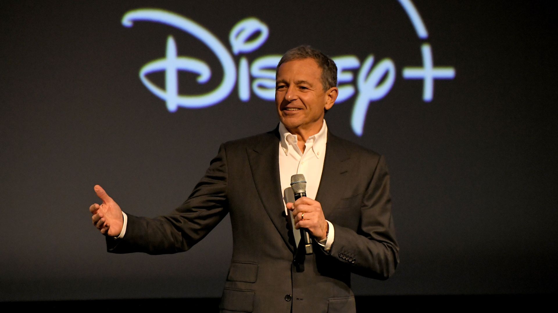 A man in a dark suit holds a microphone and speaks with the Disney logo on a screen behind him