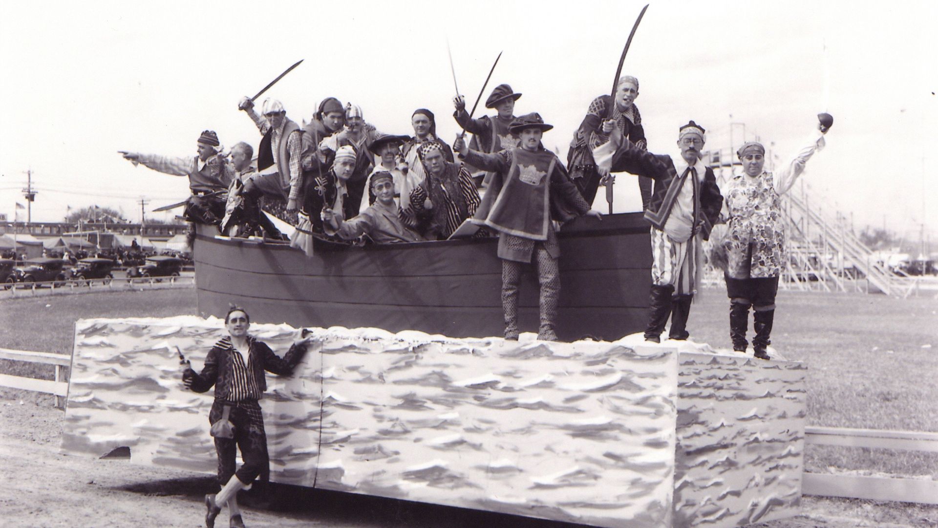 Black and white photo of men dressed as pirates with swords on a ship float decorated with waves, at an outdoor event with bleachers and parked cars in the background.