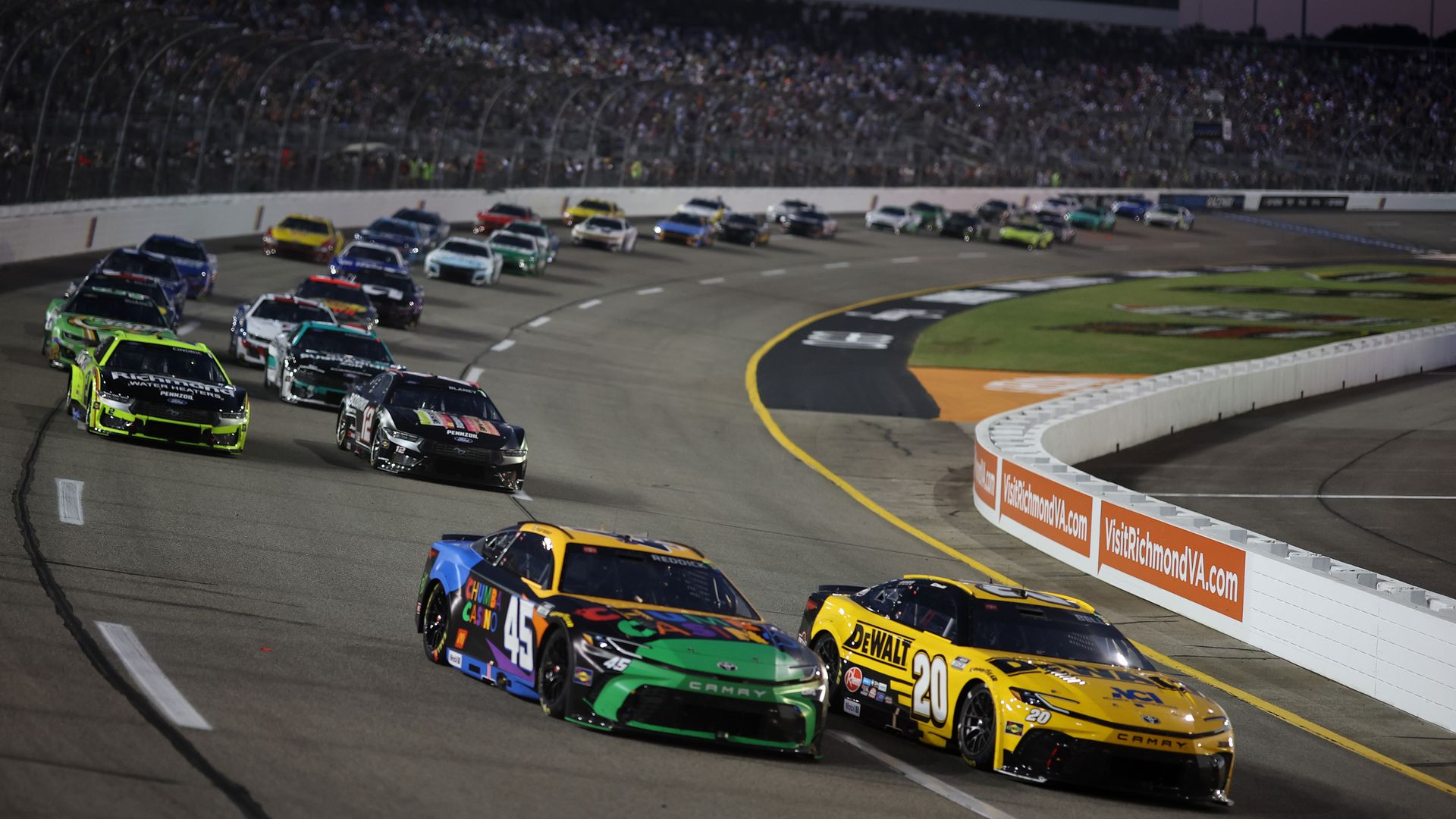 Nighttime NASCAR race on a banked oval; packed grandstands overlook a wall of orange sponsor banners VisitRichmondVA.com as colorful stock cars race along the curve.