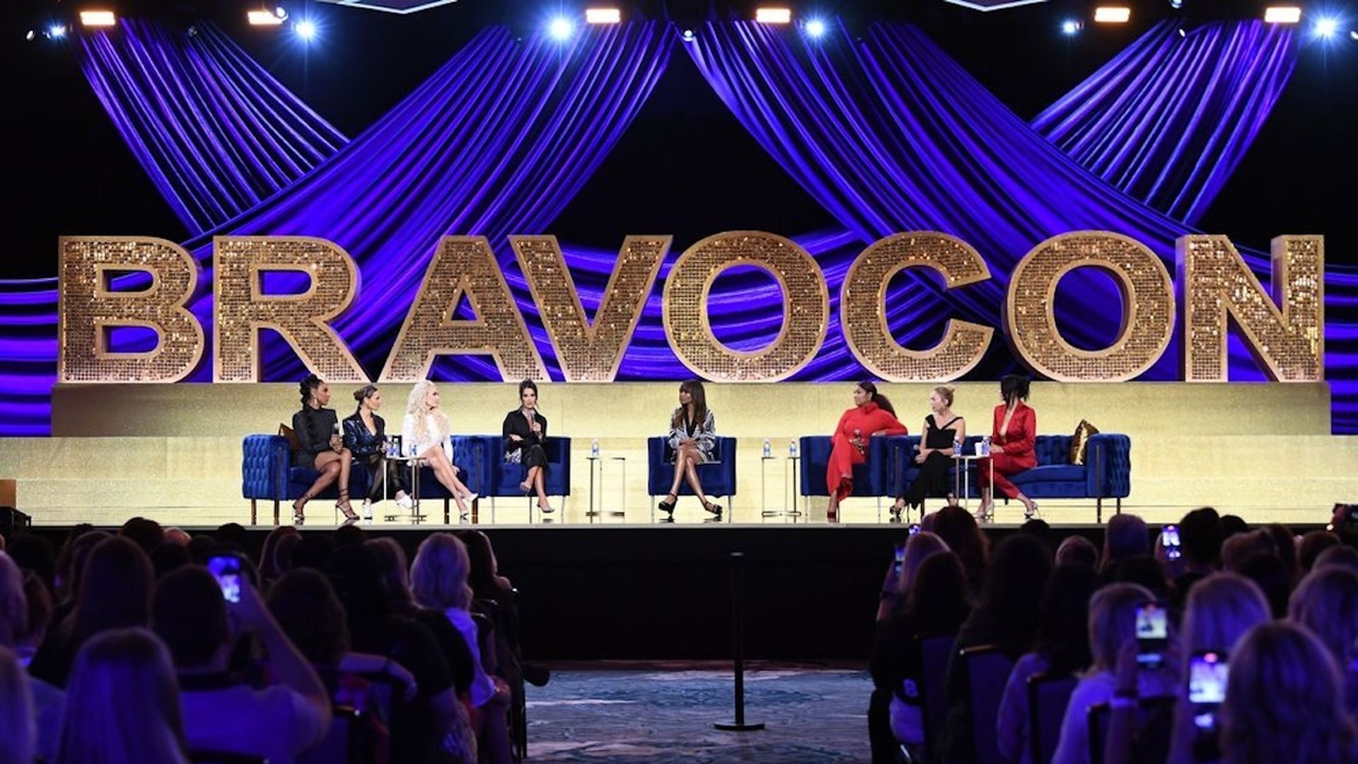 Panel of eight women seated on blue chairs on stage under large gold letters spelling BRAVOCON with blue drapes and bright stage lights, audience visible in foreground.