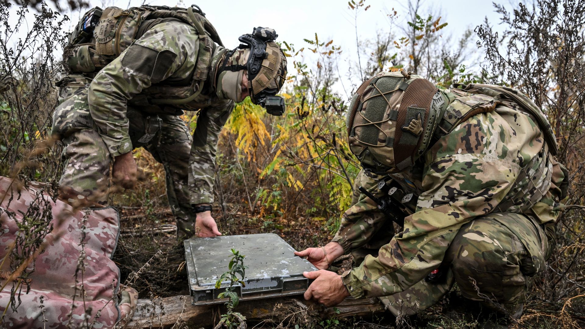 Soldiers in Ukraine, bent over in thick brush, set up a Starlink terminal. Their faces are obscured.