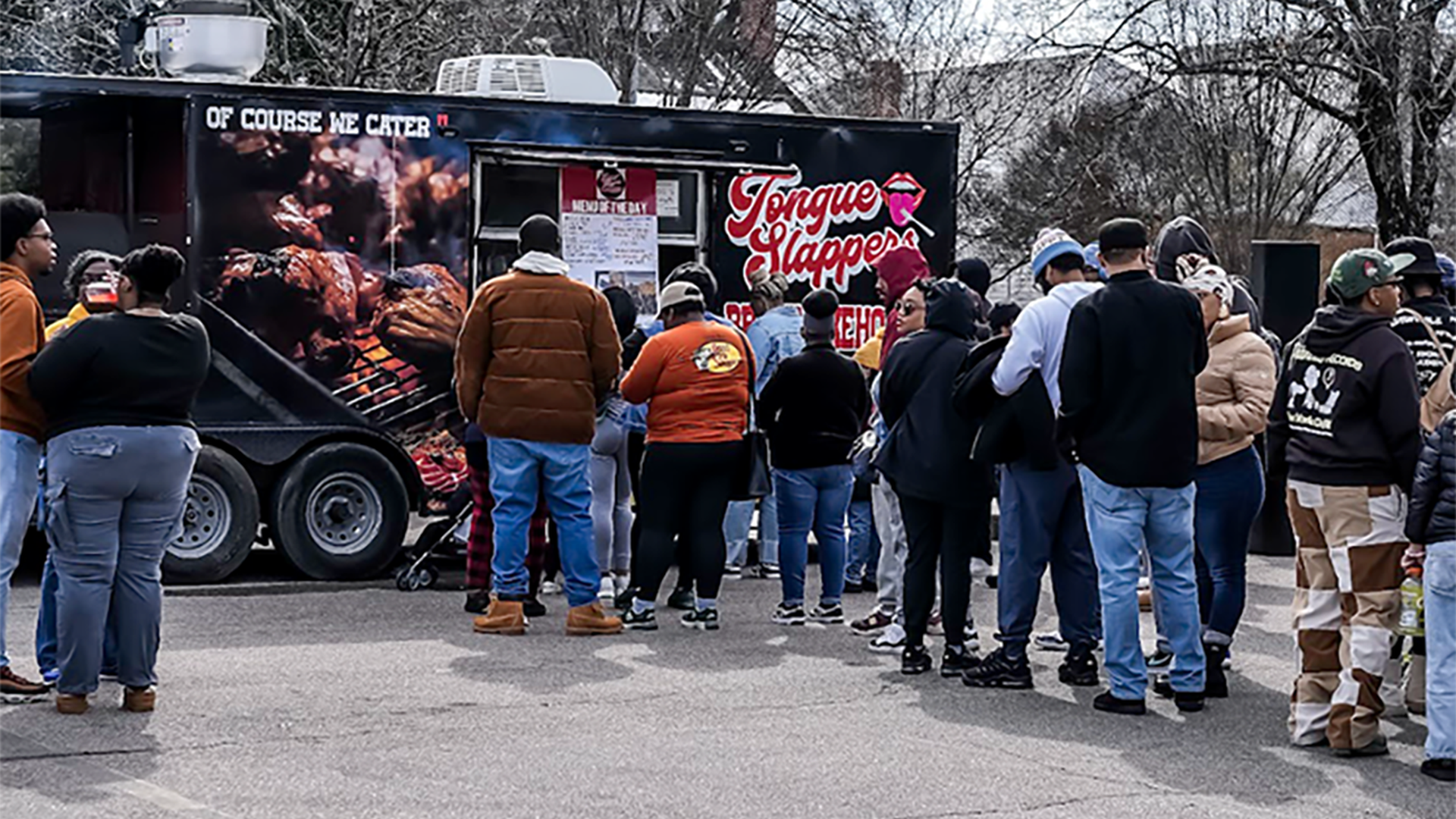 Uptown food truck rodeo. People lined up outdoors in front of Tongue Slappers BBQ food truck on a cool day with bare trees in the background.