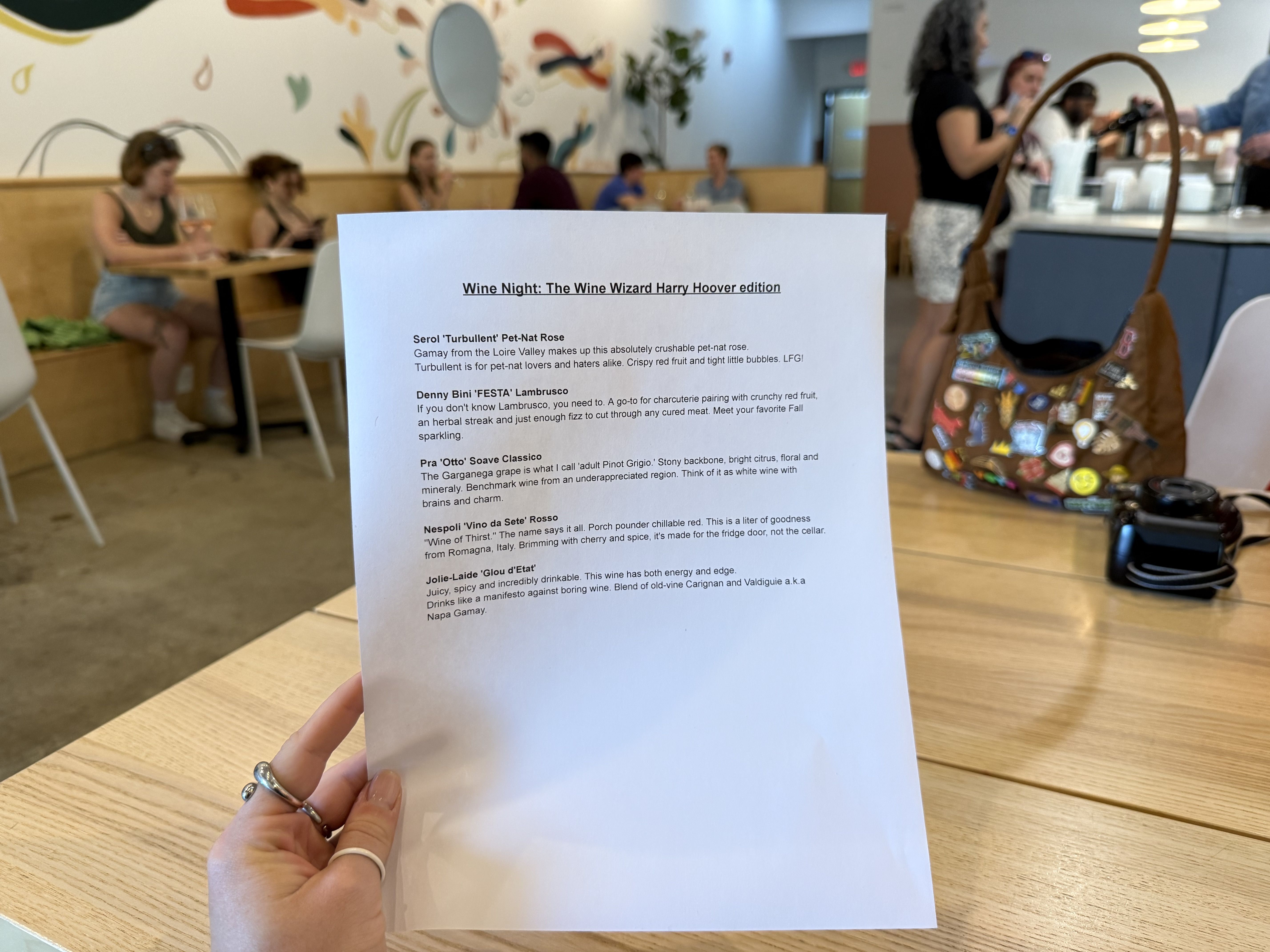 A hand holds a printed wine list titled "Wine Night: The Wine Wizard Harry Hoover edition" with descriptions of five wines, in a bright, modern cafe with people and a colorful bag in the background.