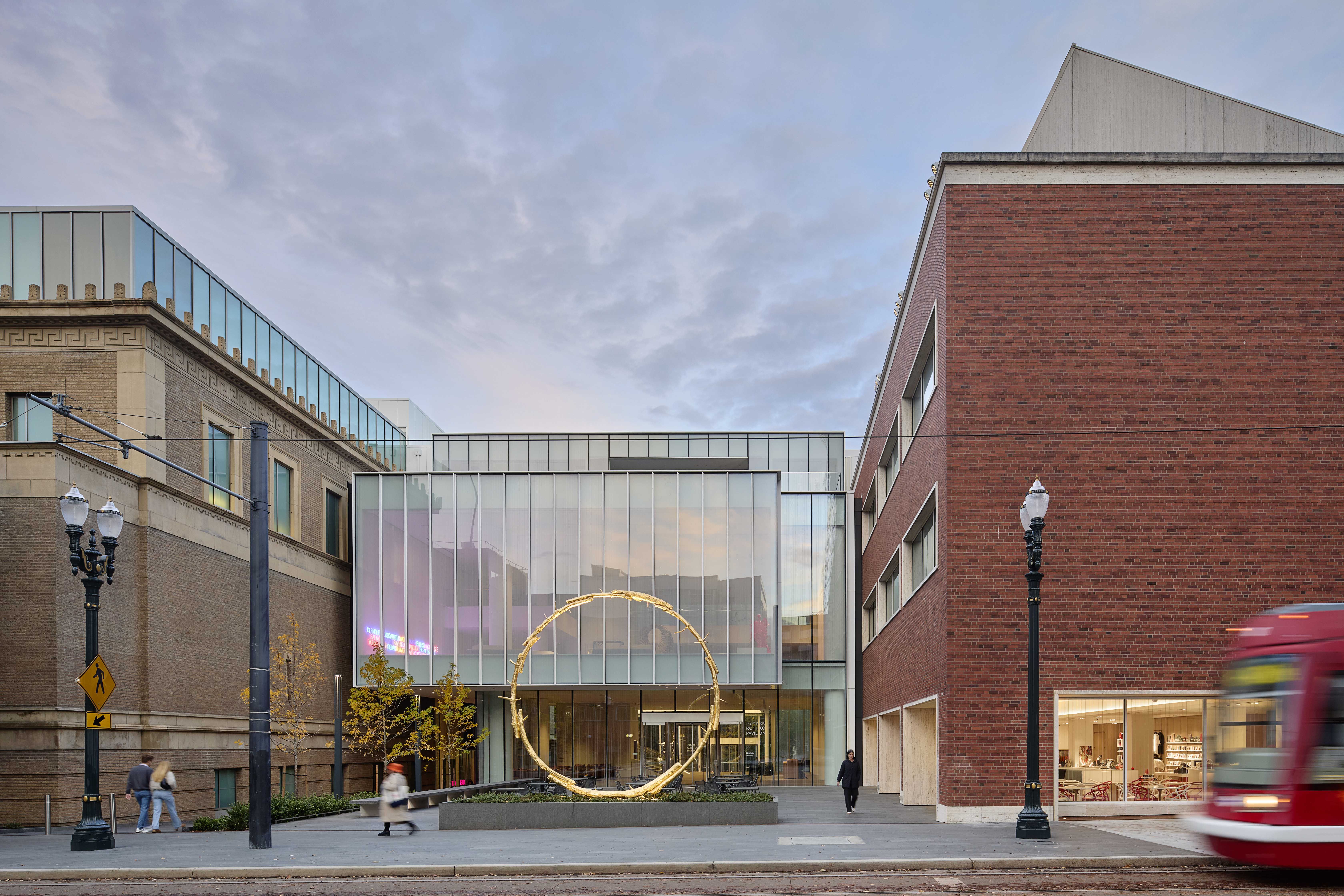Urban street scene with modern glass-front building featuring large golden circular sculpture, flanked by brick buildings, pedestrians walking, and a red streetcar passing by.