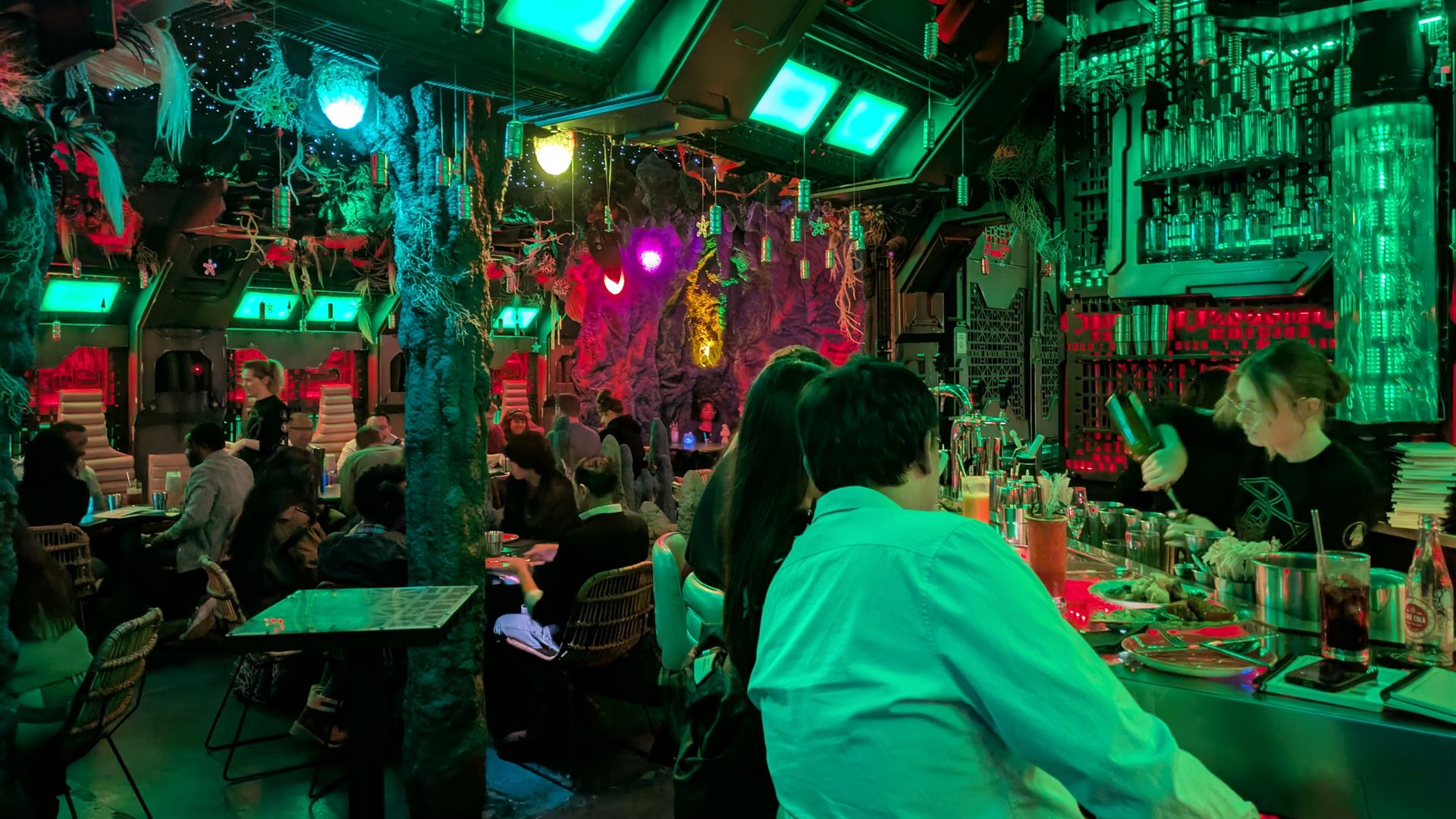 Crowded futuristic bar with green and purple neon lights, people sitting and chatting, bartender mixing drinks, and sci-fi decor including hanging lights and textured walls.