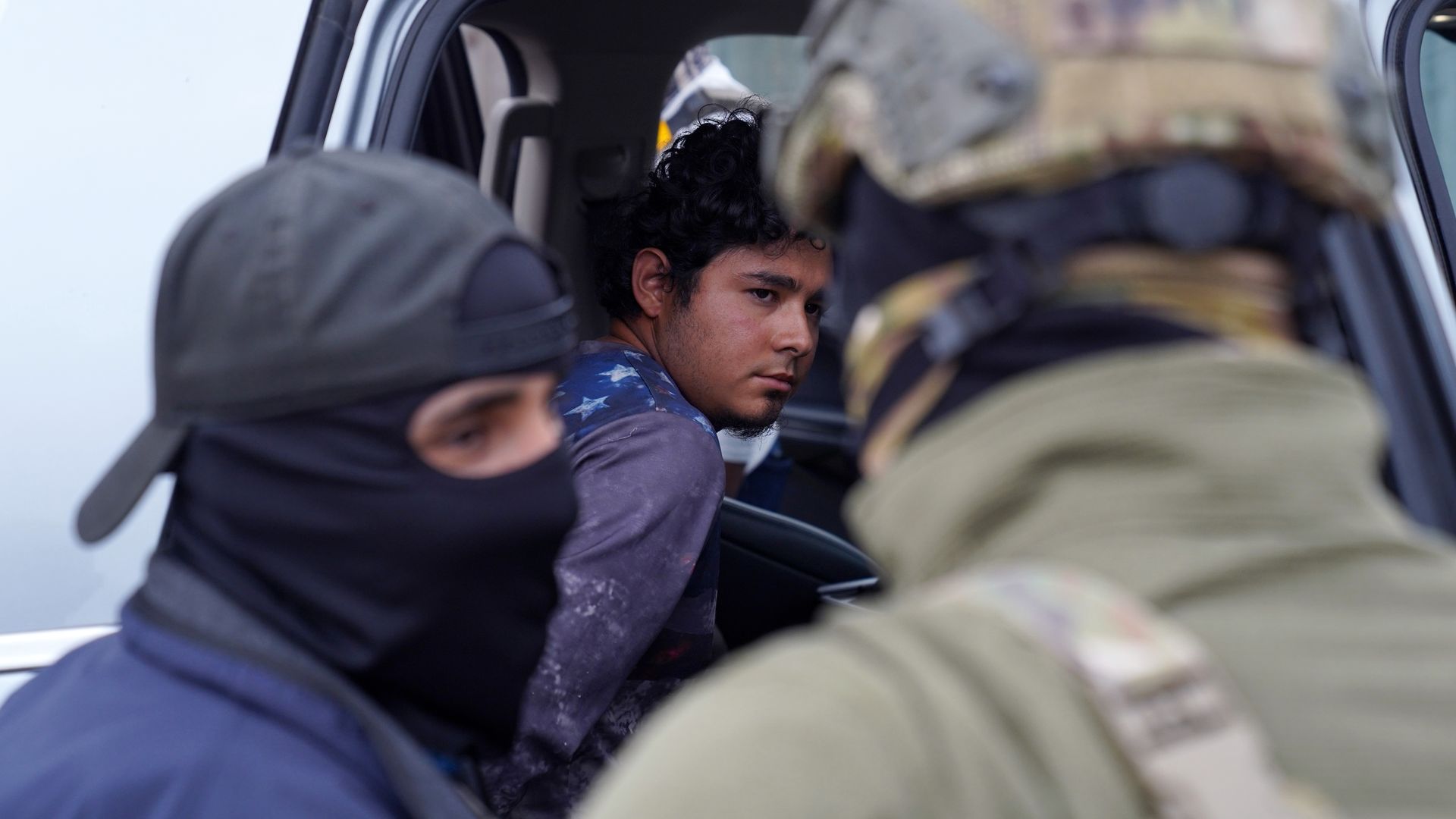 A man sits in the back of an SUV, viewed through an open door. Two agents, one of them masked, are seen out of focus in the foreground.