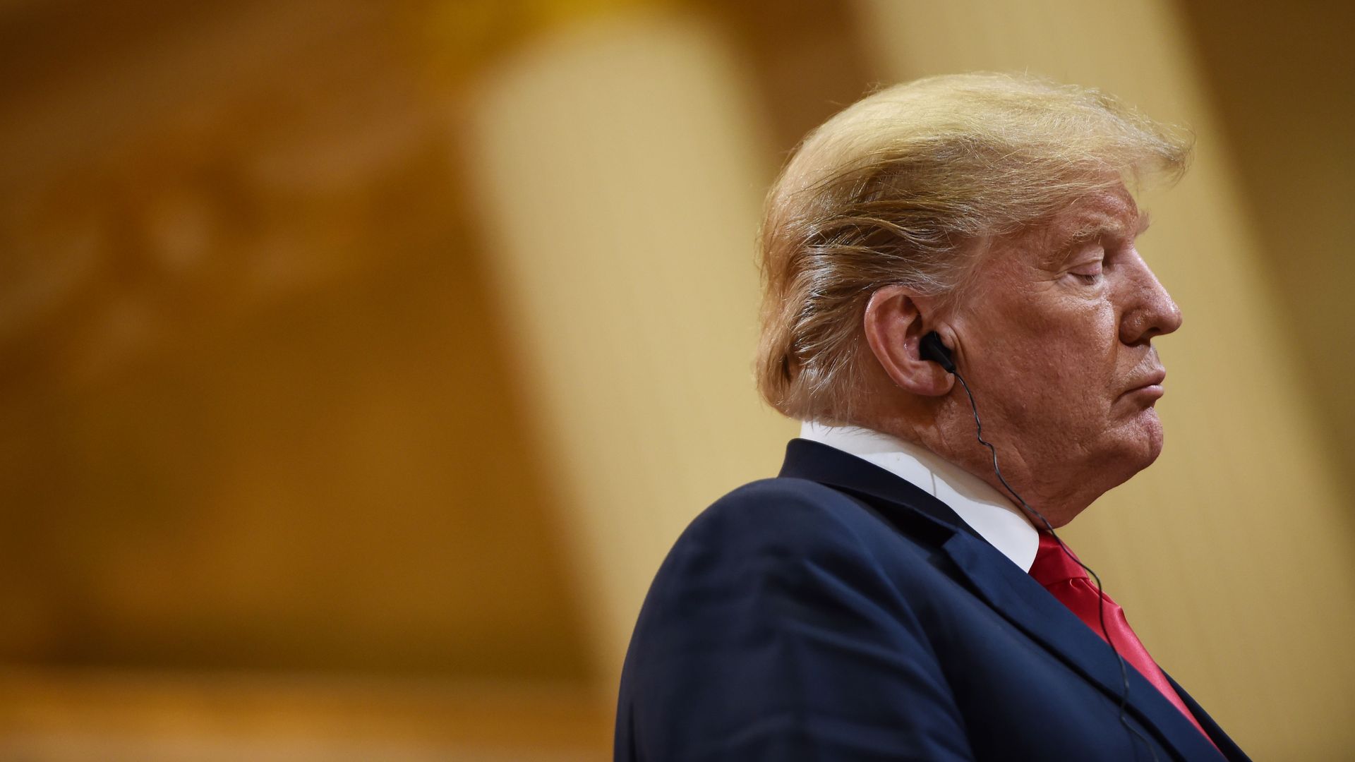 Donald Trump looking down and forlorn before a golden background.