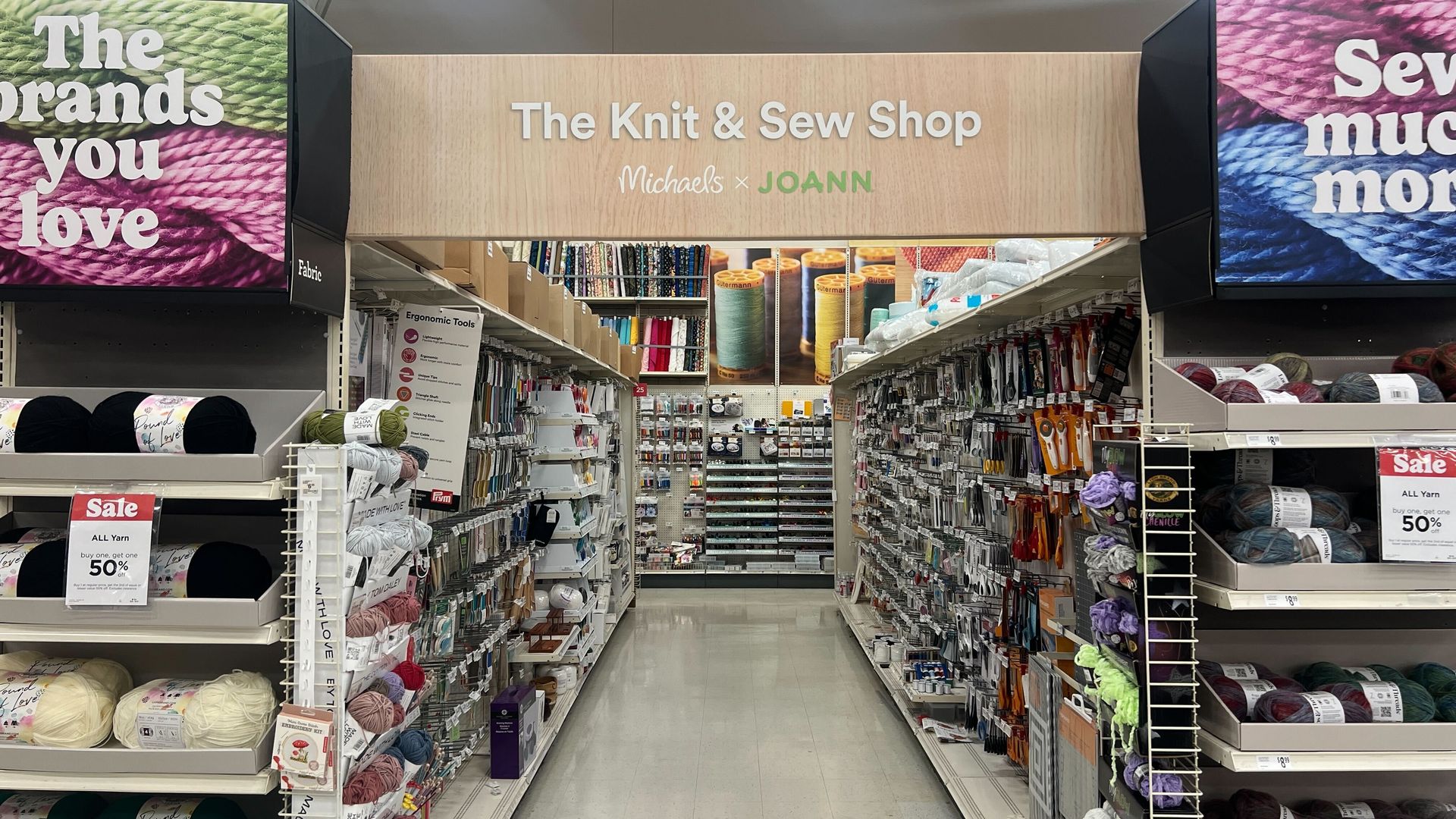 A well-stocked aisle in a craft store labeled The Knit & Sew Shop, featuring yarn in various colors on shelves, sewing tools, and accessories on both sides, with promotional sale signs.