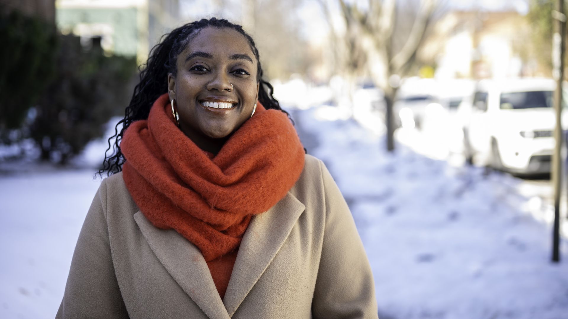 Doni Crawford is photographed smiling and wearing a beige coat and bright red scarf standing outside on a snowy street on a sunny day.