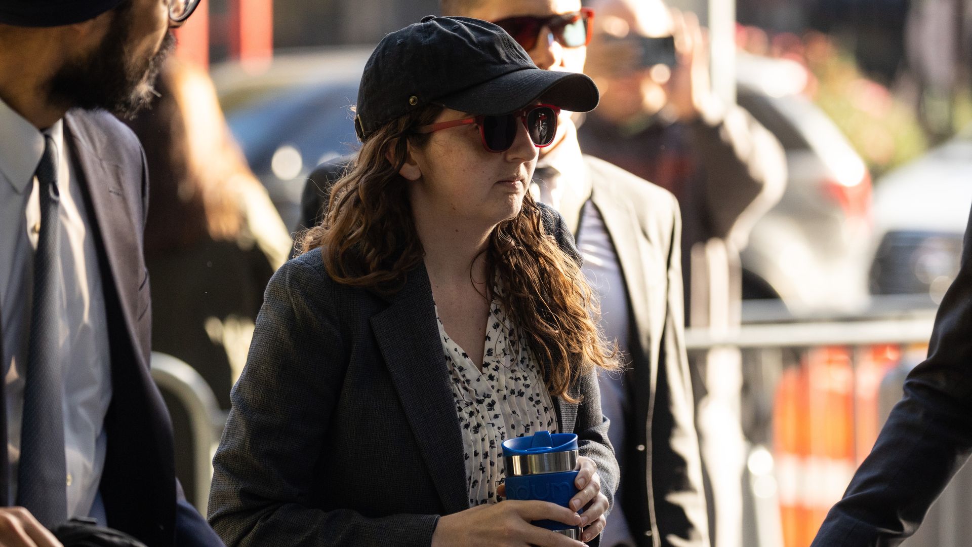 A woman with sunglasses and a had pulled tight on her head holds a blue mug, with security around her.