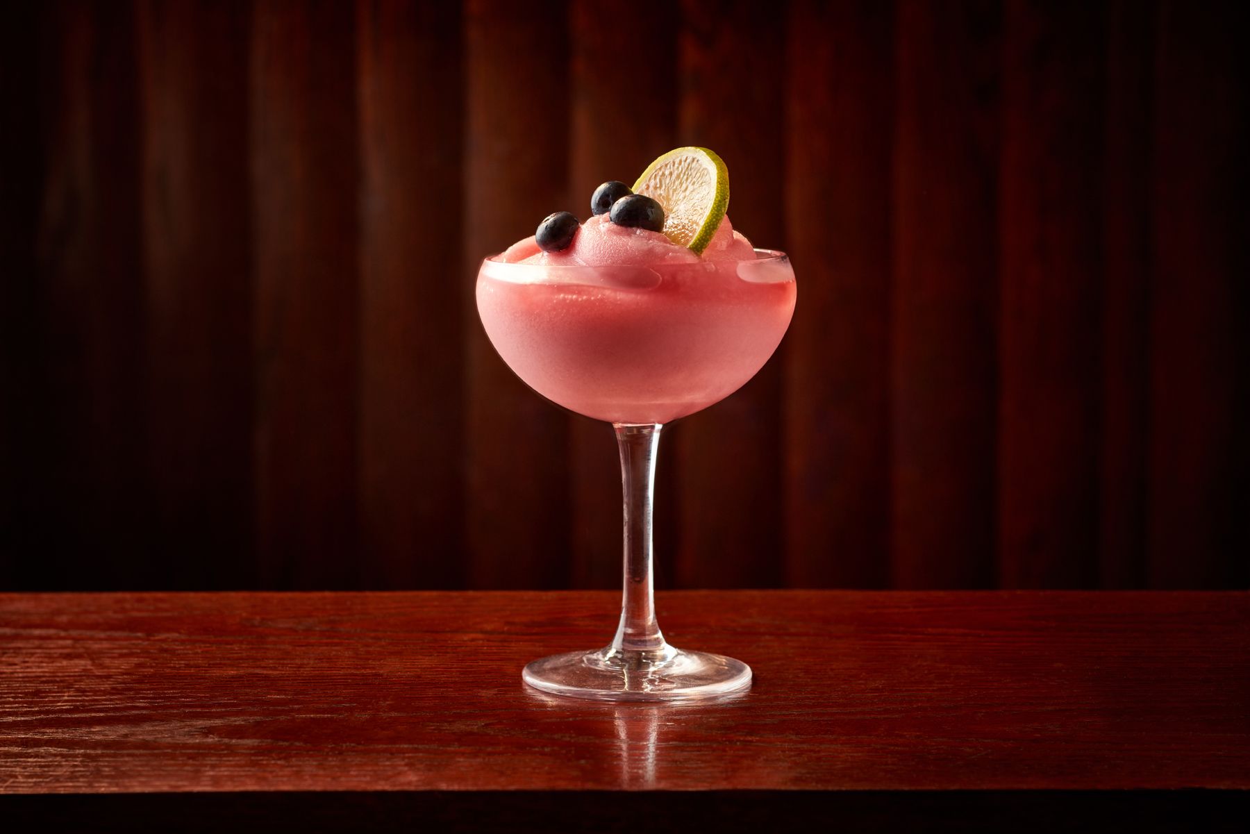 Pink frozen cocktail in a coupe glass garnished with three blueberries and a lime slice on a wooden surface with a dark wood background.