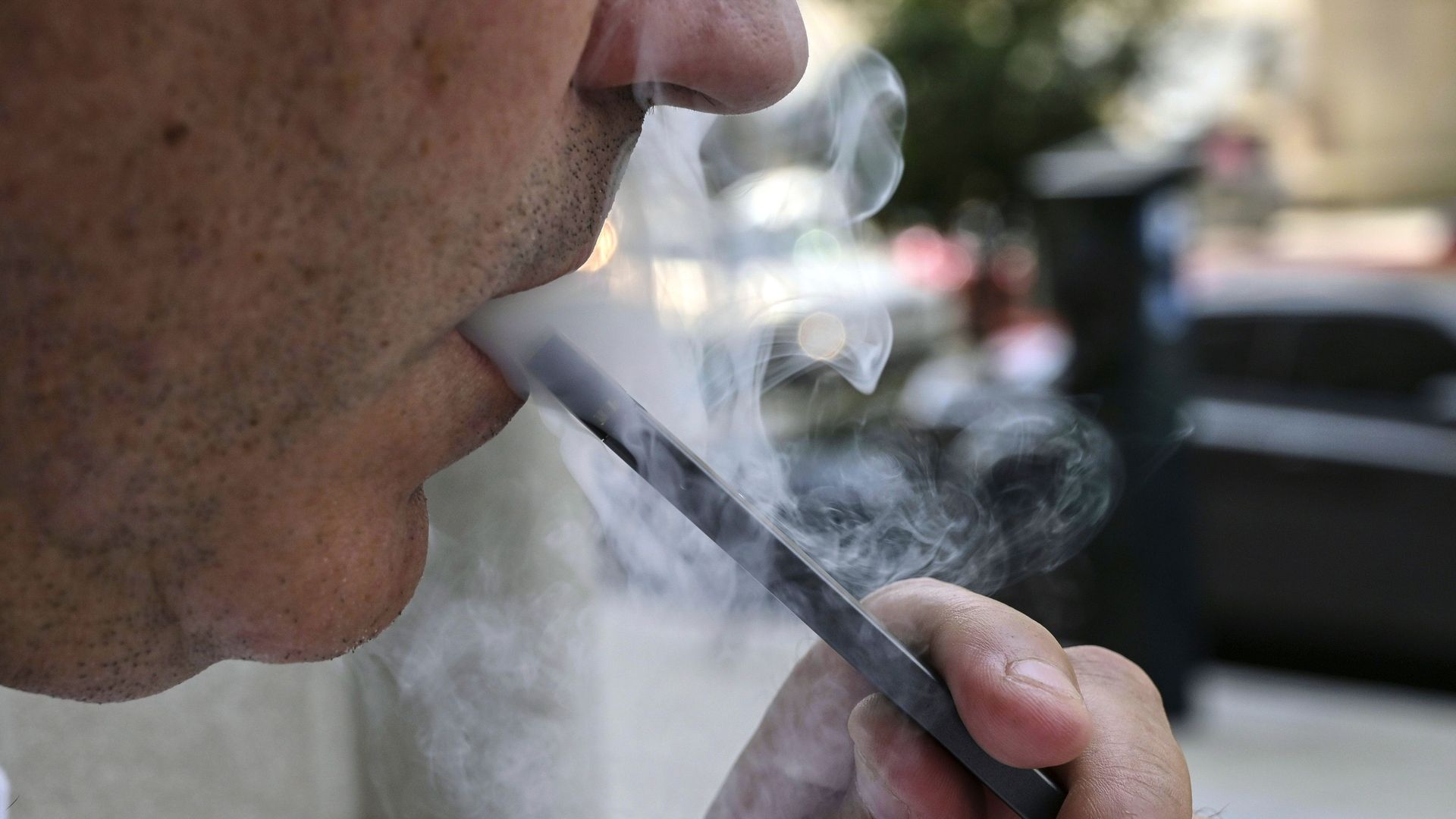 In this image, a man blows a cloud of smoke while vaping outside