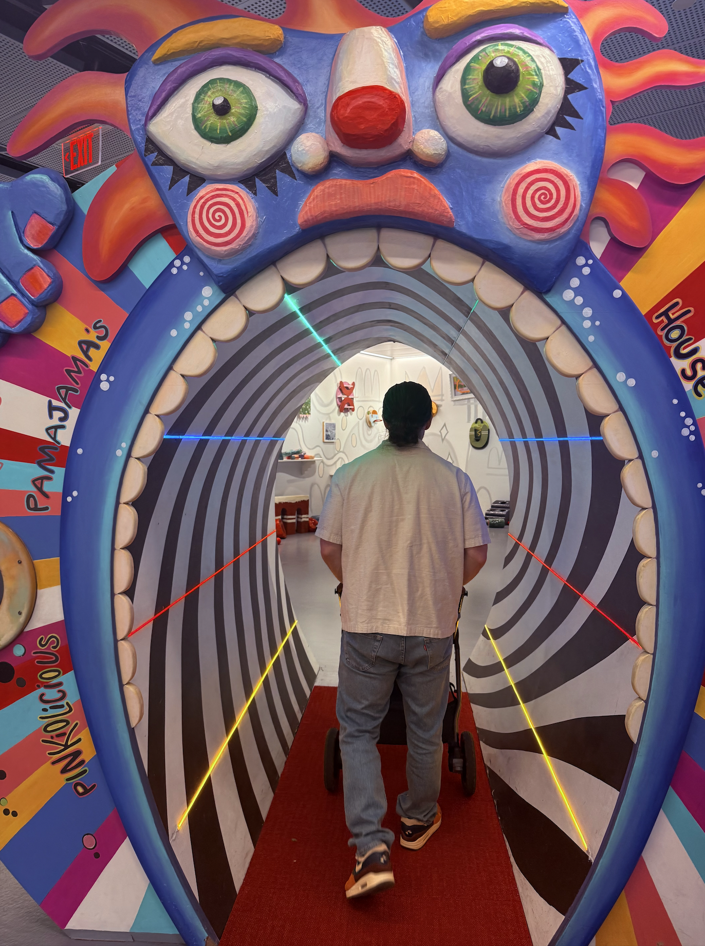 Inside a vivid art installation, a person walks into a tunnel formed by a giant blue monster face with green eyes, red nose, spiraled cheeks, orange flames, and black-and-white striped inner walls.