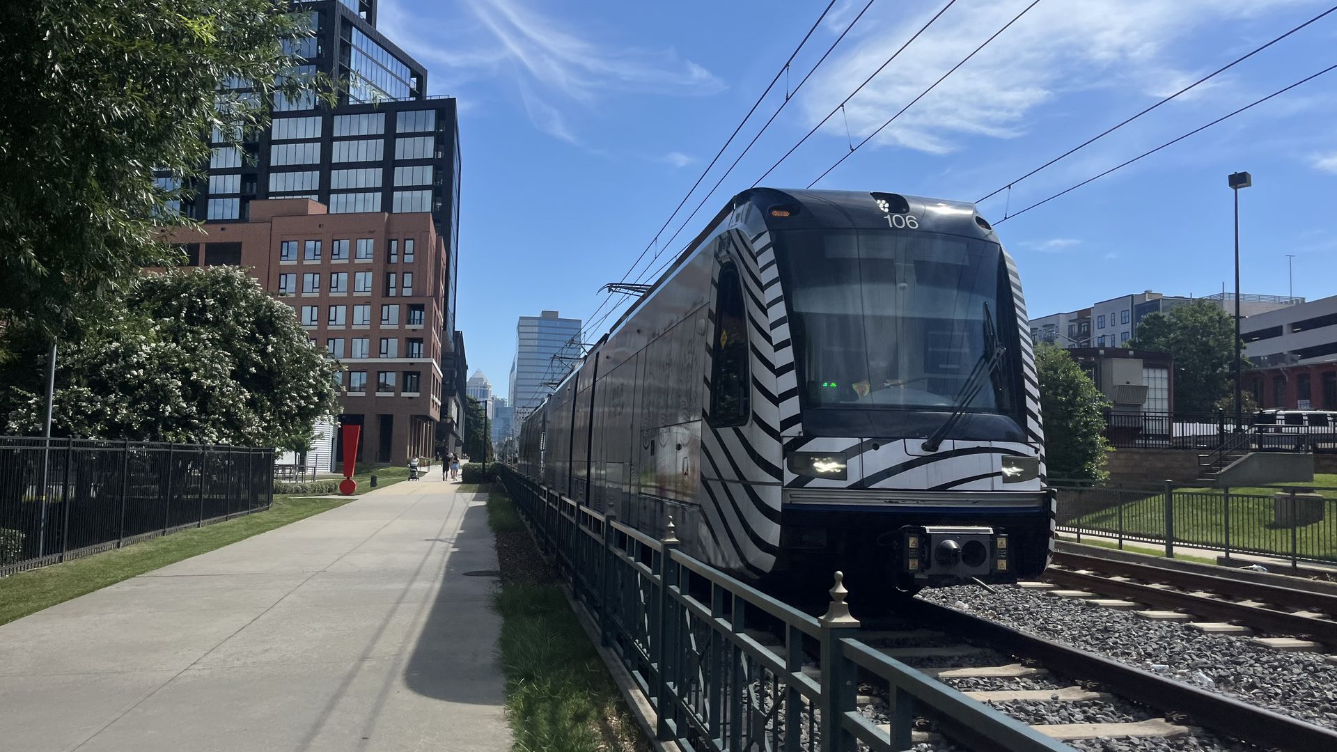 Black and white striped light rail train 106 on tracks next to a sidewalk in a sunny urban area with tall buildings and blue sky.