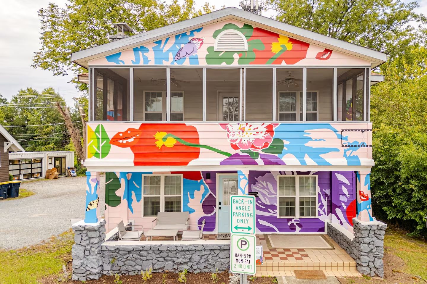 Two-story house with colorful floral mural featuring red, purple, green, blue, and yellow flowers. Lower porch has seating; sign in front restricts parking to back-in angle, 8am-5pm.