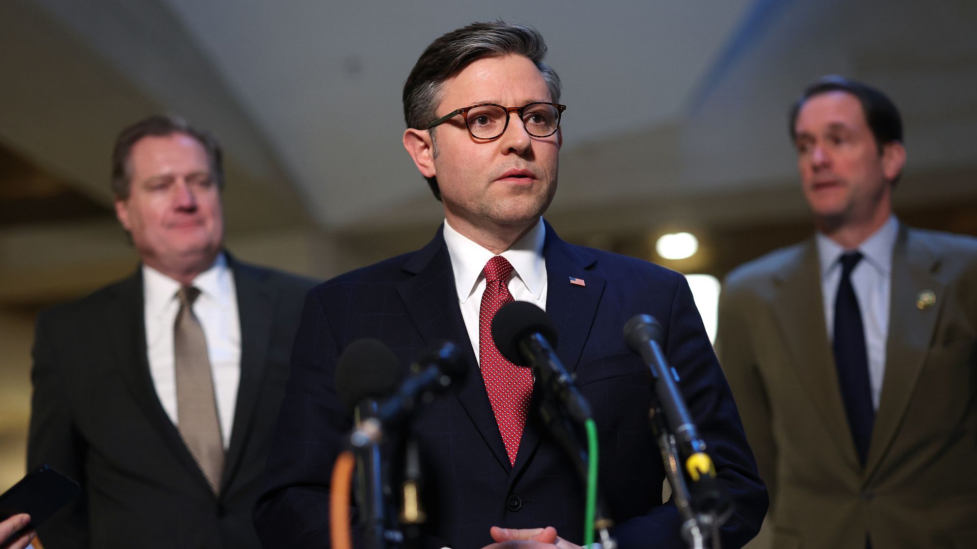 Speaker Mike Johnson, wearing a blue suit, white shirt, red tie and glasses, stands in front of microphones flanked by Reps. Mike Turner and Jim Himes.