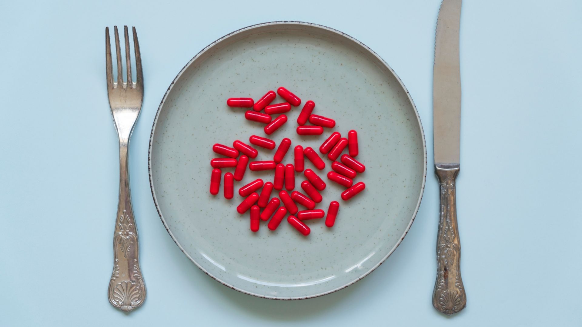 A speckled ceramic plate on a pale blue background holds a cluster of bright red capsules; a vintage fork lies to the left and a serrated knife to the right.