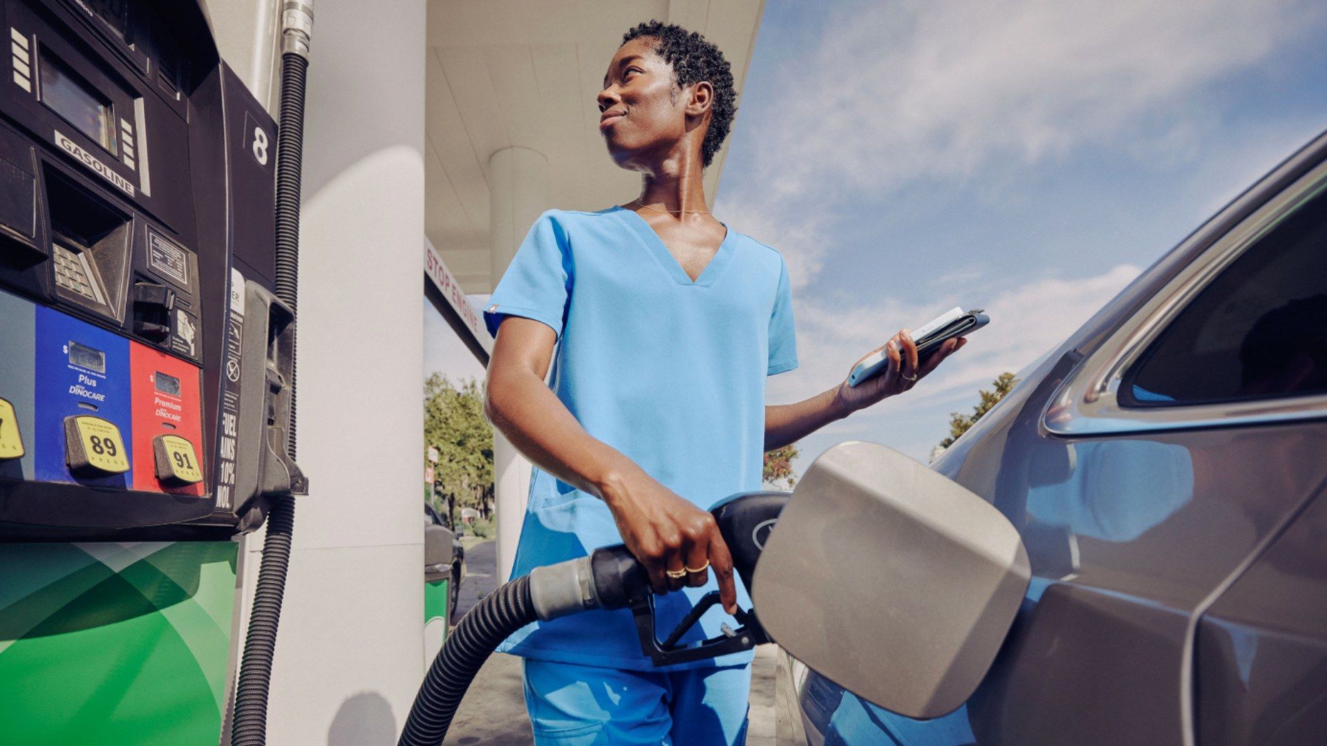 A person in medical scrubs fills their car with gas at a pump while holding a phone and looking off into the distance.