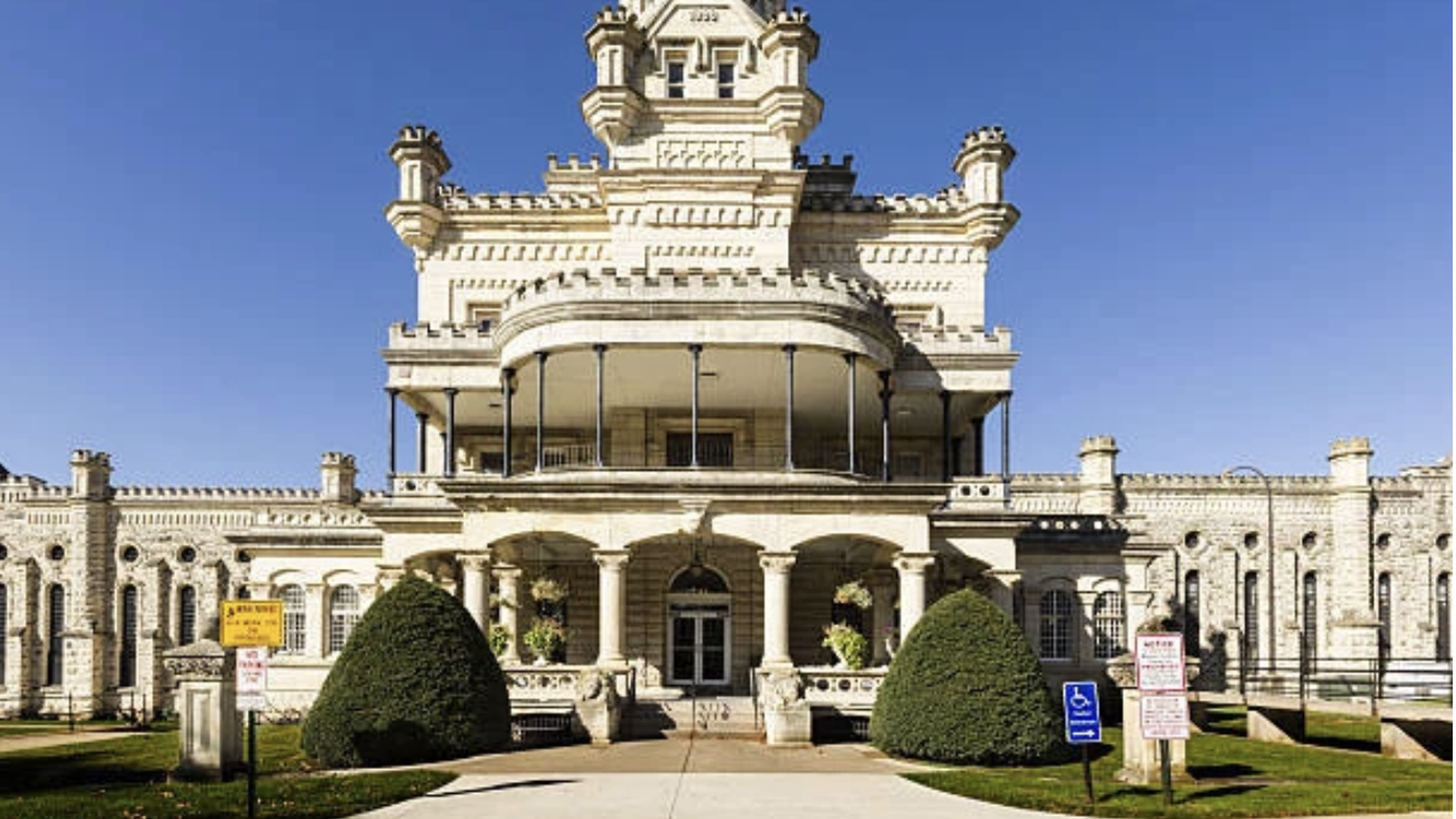 White, castle-like building with a tall, ornate central tower, curved balcony, and arched entrance; manicured shrubs and a stone wall frame the facade under a clear blue sky.