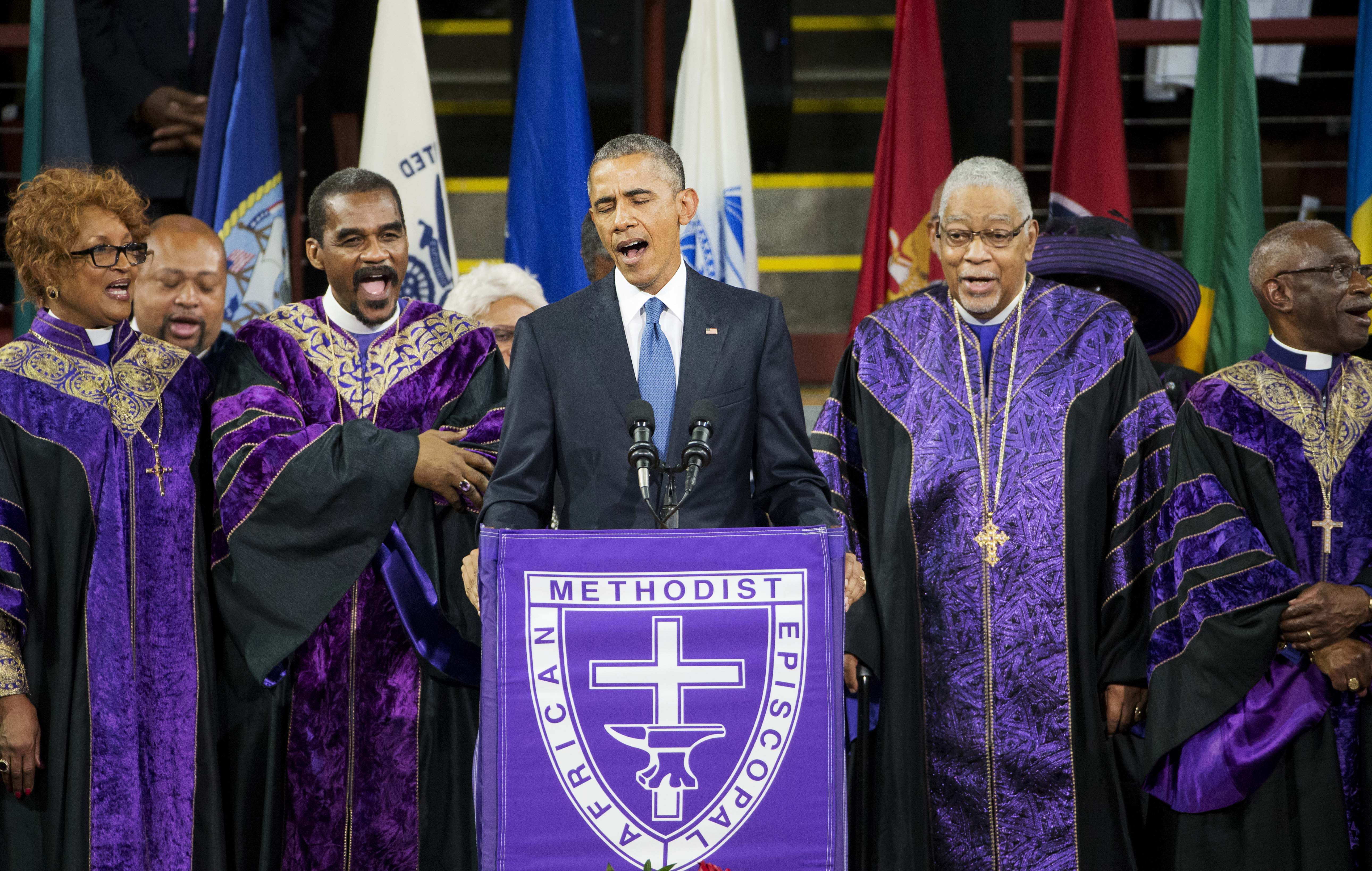 A man stands at a podium covered with a purple cloth featuring a cross and the words 'African Methodist Episcopal.' He is surrounded by a choir wearing purple and gold church robes. The group appears to be singing together. Various flags are visible in the background.