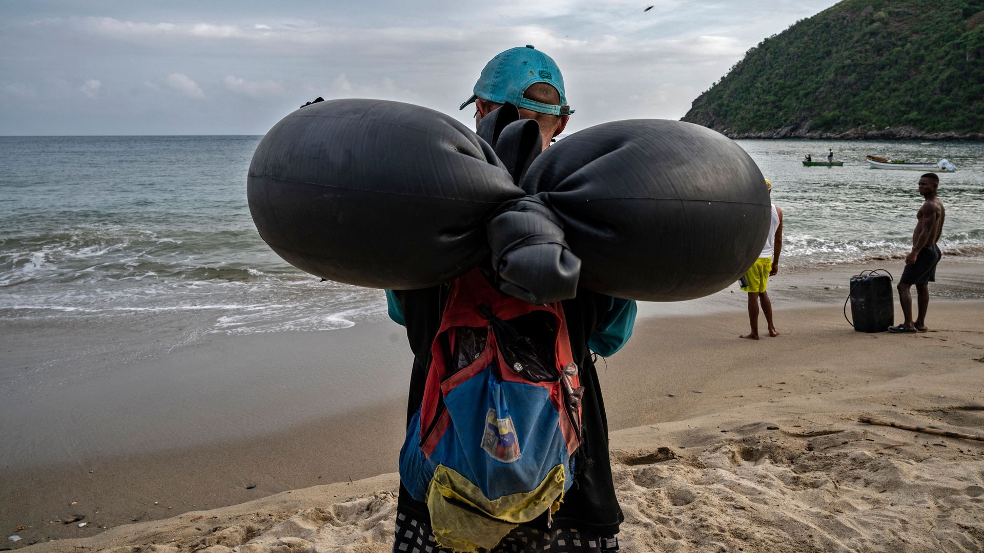 A man wearing a baseball cap, black t-shirt and backpack carries a black inflatable tube on his back as he walks toward the ocean on the sand. 
