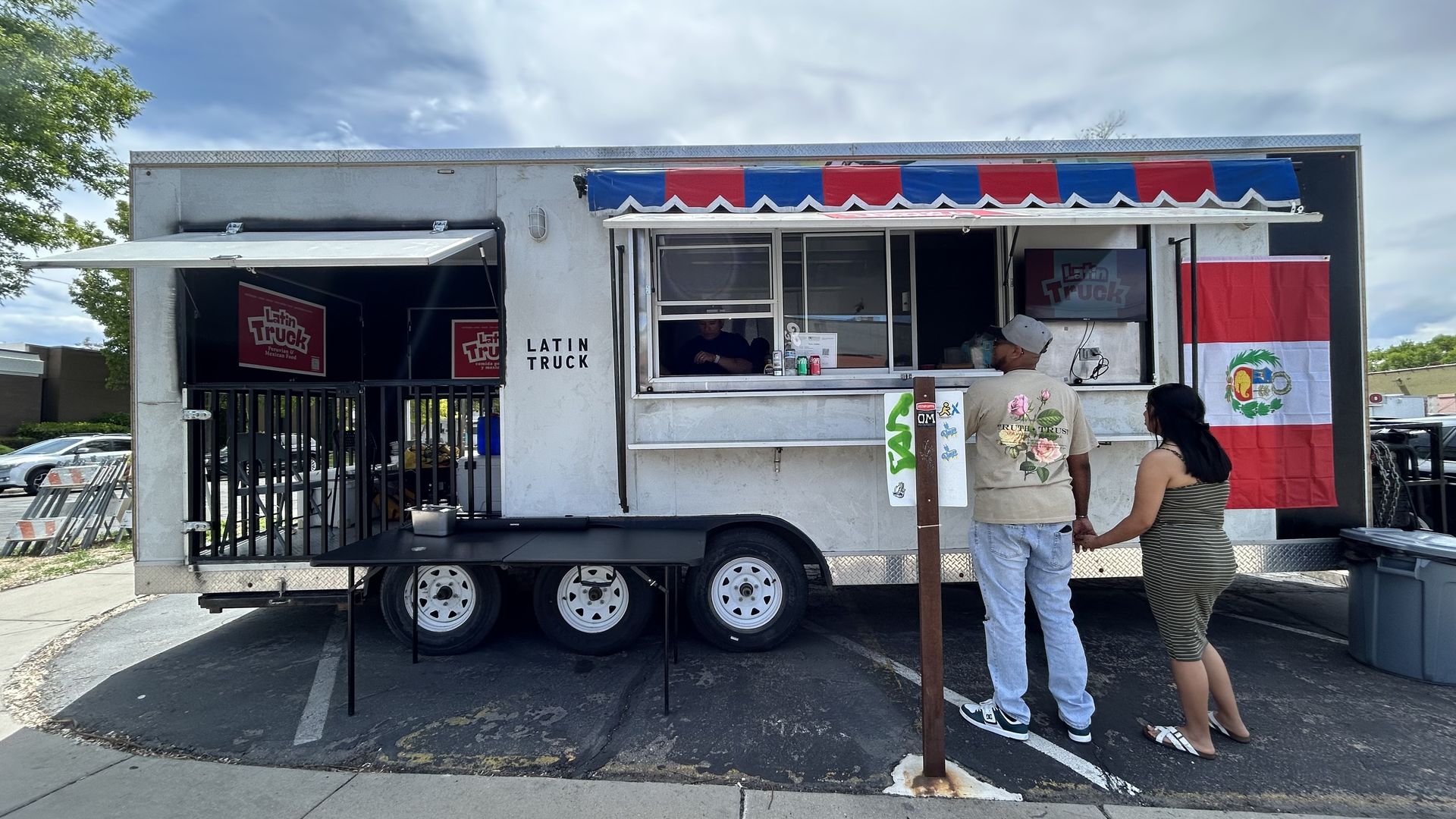 White Latin Truck food trailer with a red-blue striped awning and open service window. A man and a woman stand in line; a Peru flag hangs on the side; blue sky and trees in the background.