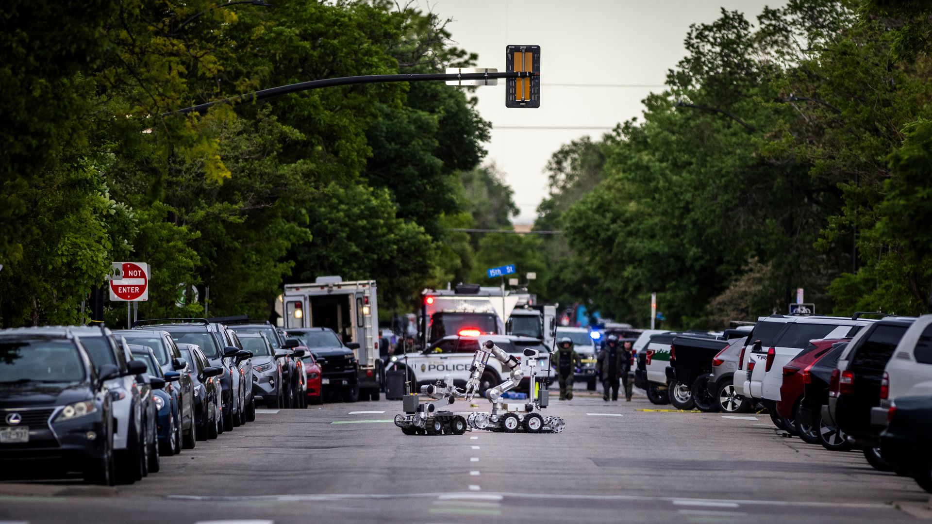 A street is seen with cars parked on either side and trees surrounding them. A robot is seen in the middle 