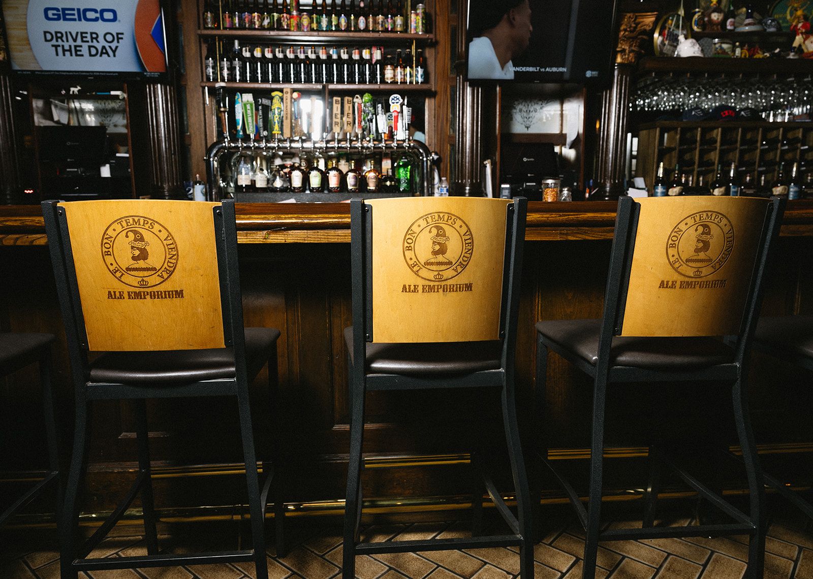 Three wooden-backed bar stools with the "Ale Emporium" logo face a polished bar; behind them are beer taps and bottle shelves, with TVs and dark wood decor.