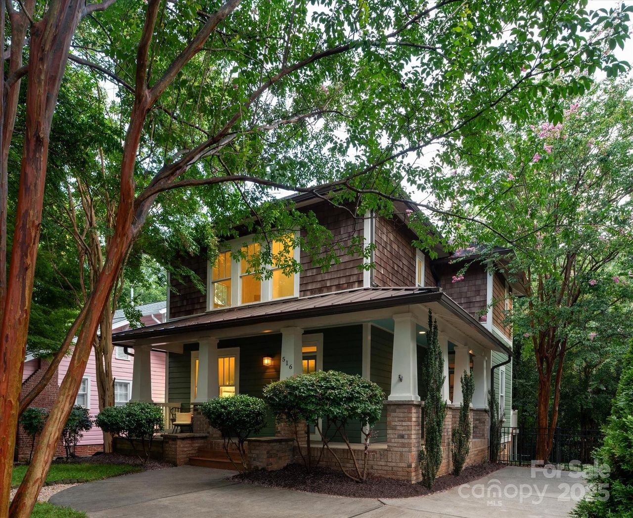 Two-story house with green siding and brown shingles, lit windows, front porch with white columns, surrounded by green trees and bushes, driveway in front.