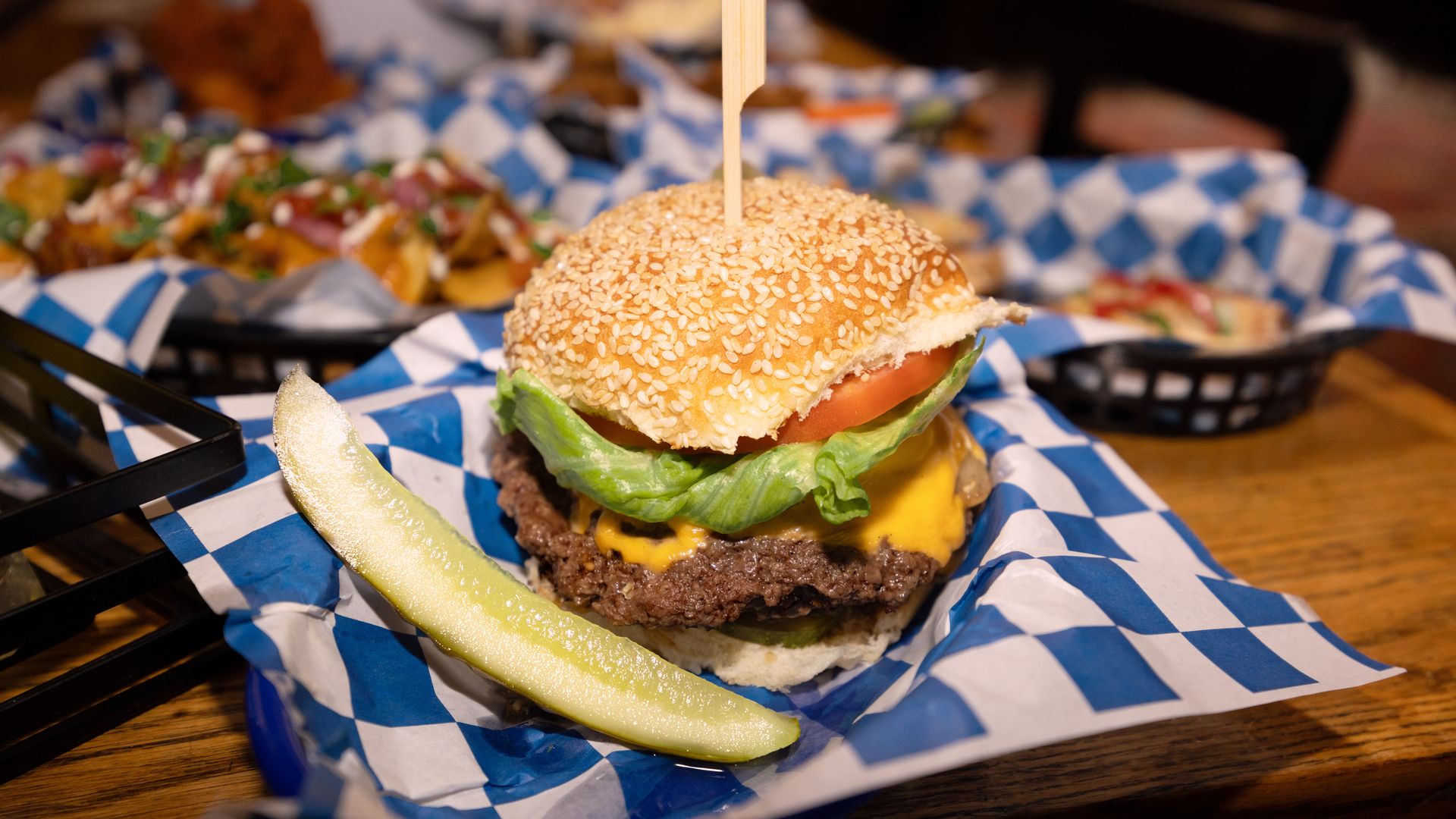 Cheeseburger with lettuce, tomato, and sesame seed bun served with a pickle spear on blue and white checkered paper in a basket on a wooden table.
